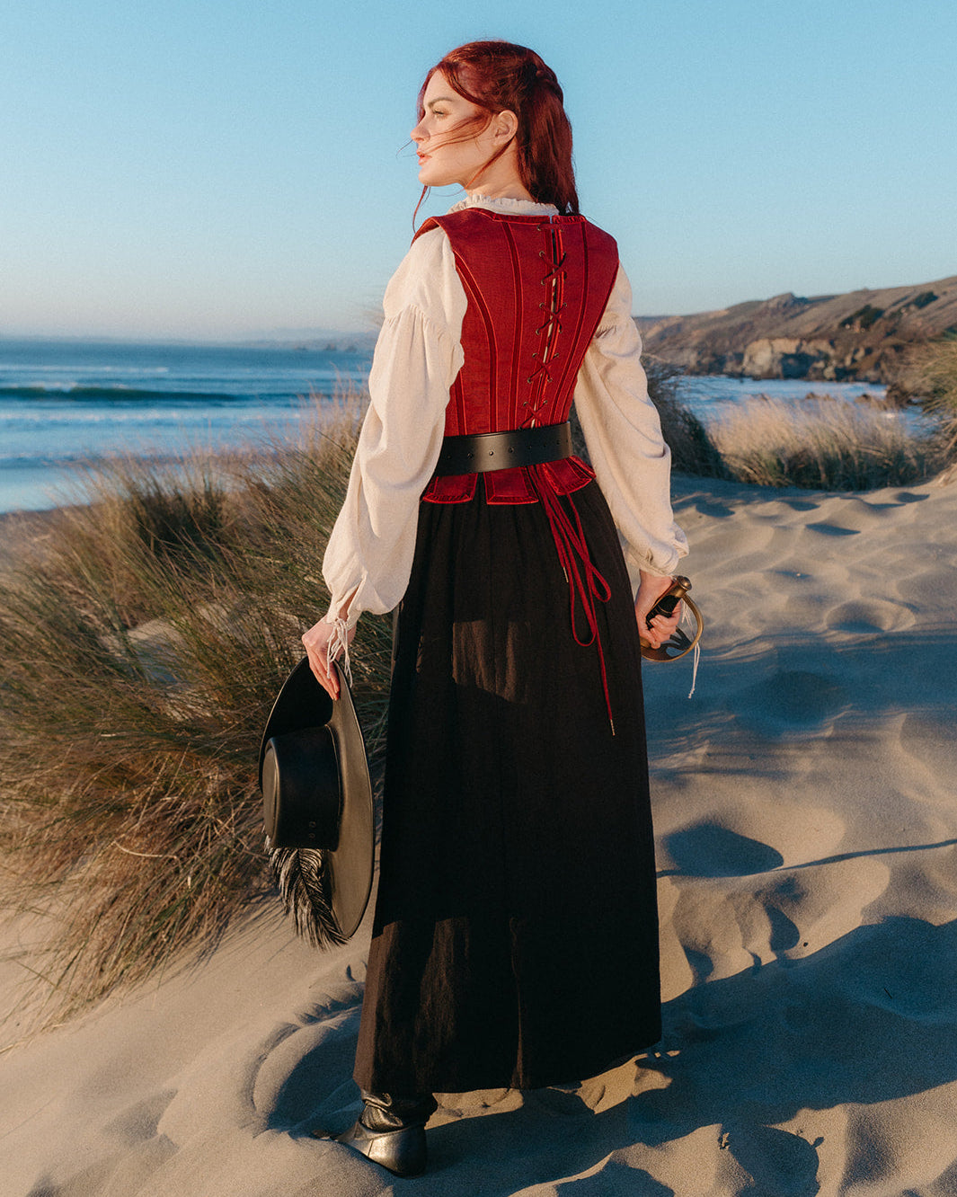 woman on beach wearing Red Pirate Doublet with white blouse black skirt holding hat and sword