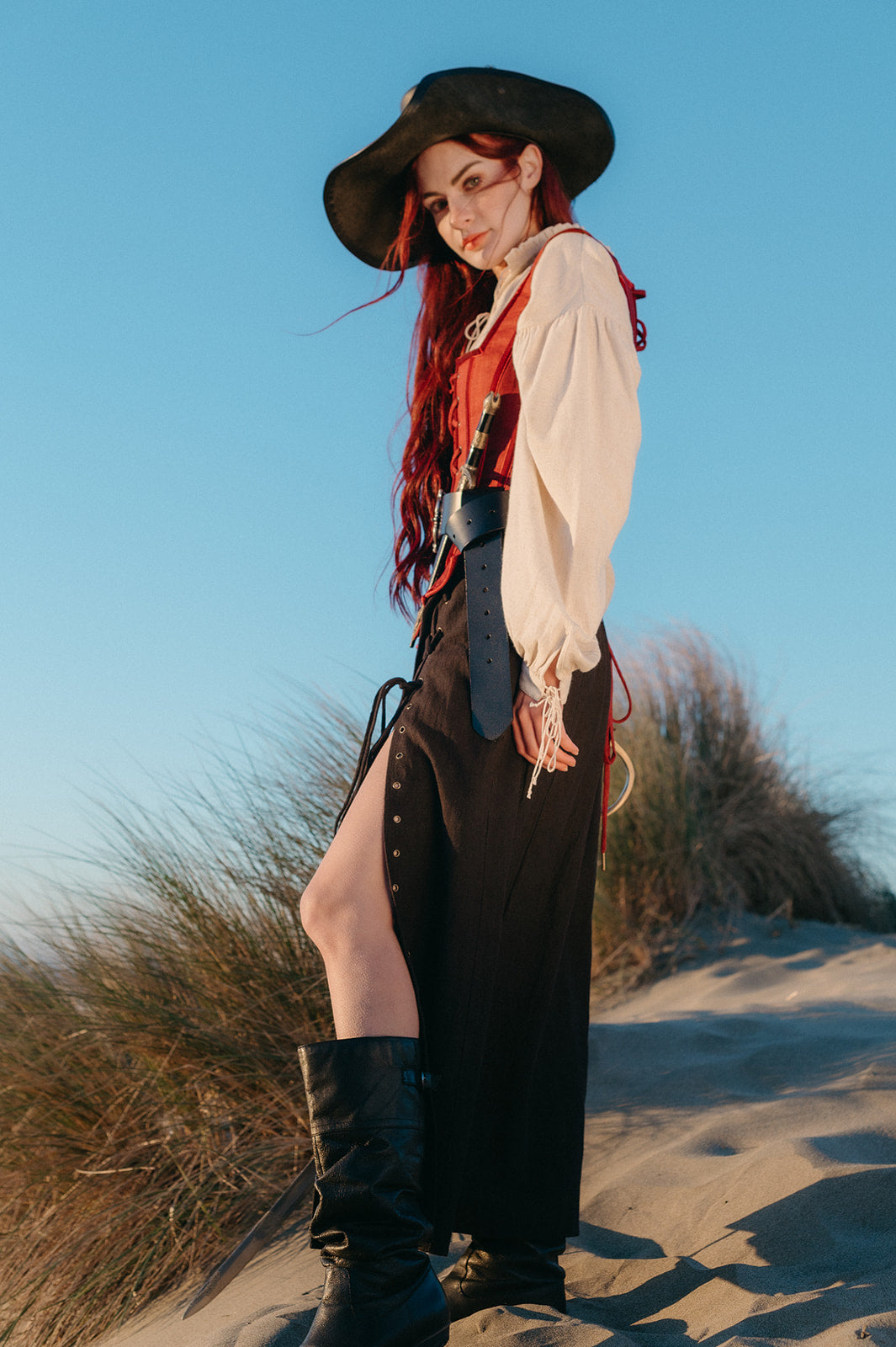 Woman wearing a Tavern Skirt with a slit, paired with boots and a medieval-inspired outfit on a sandy dune at sunset