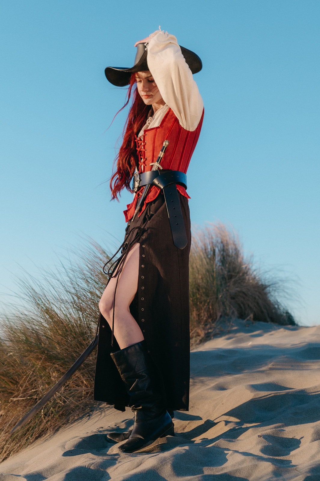 Woman wearing Leather Pirate Belt with a red corset, black skirt, and boots standing on sand dunes under clear sky