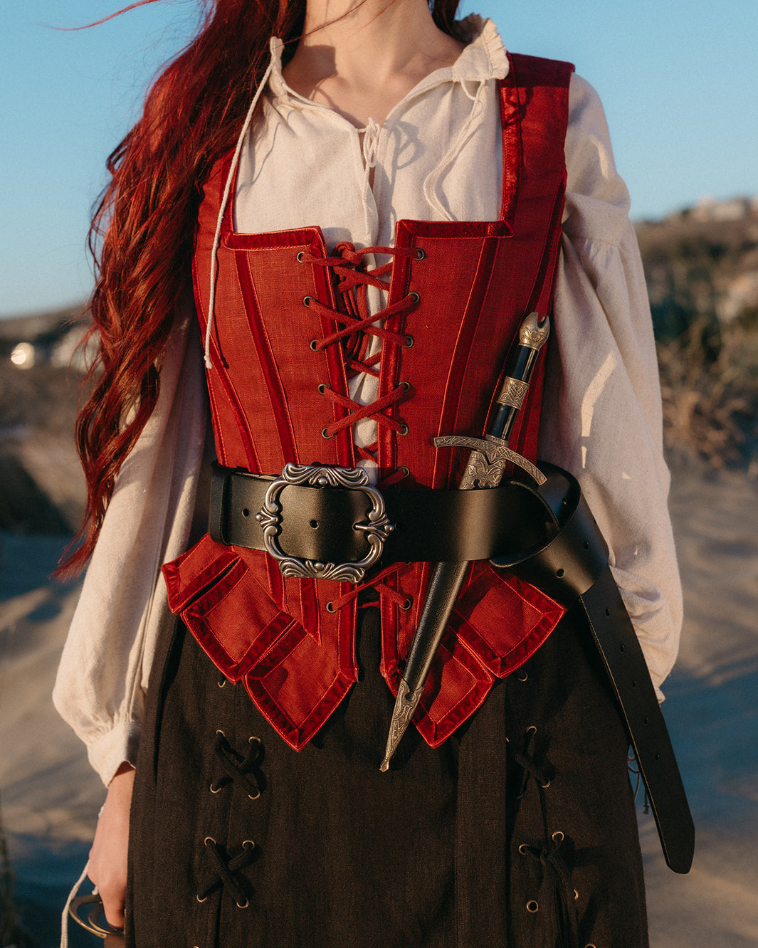 Woman wearing a red corset and a Leather Pirate Belt with an ornate buckle and attached dagger sheath outdoors