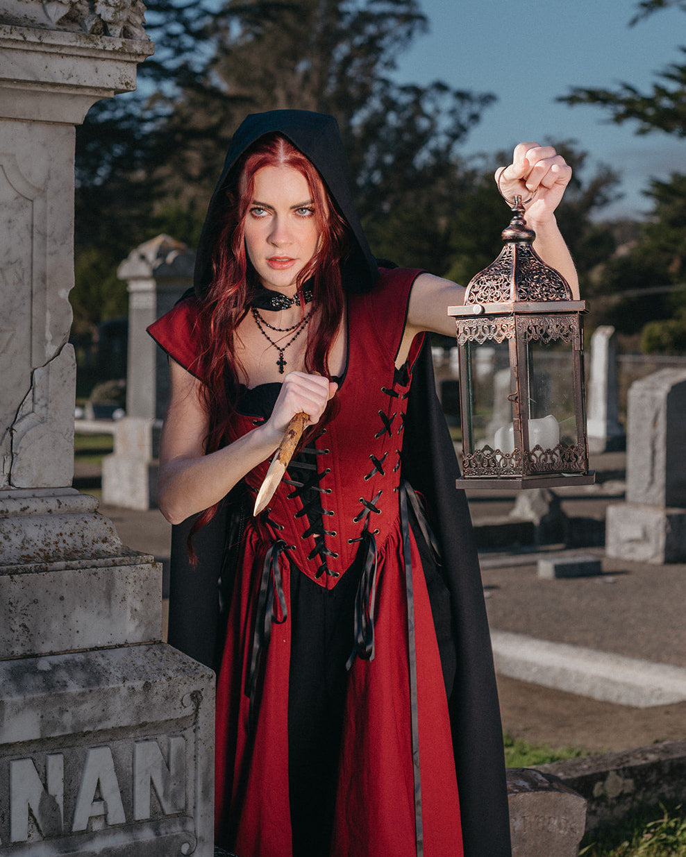 Woman in a graveyard wearing a Red Vampire Slayer Corset Dress holding a lantern and a dagger with intense expression