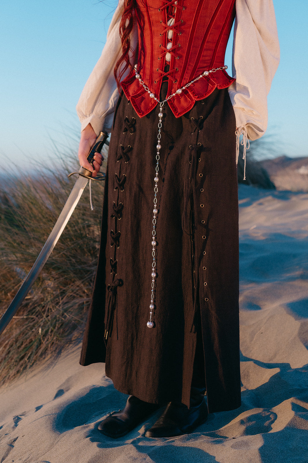 Person wearing a Tavern Skirt and holding a sword standing on sandy terrain with grass in the background