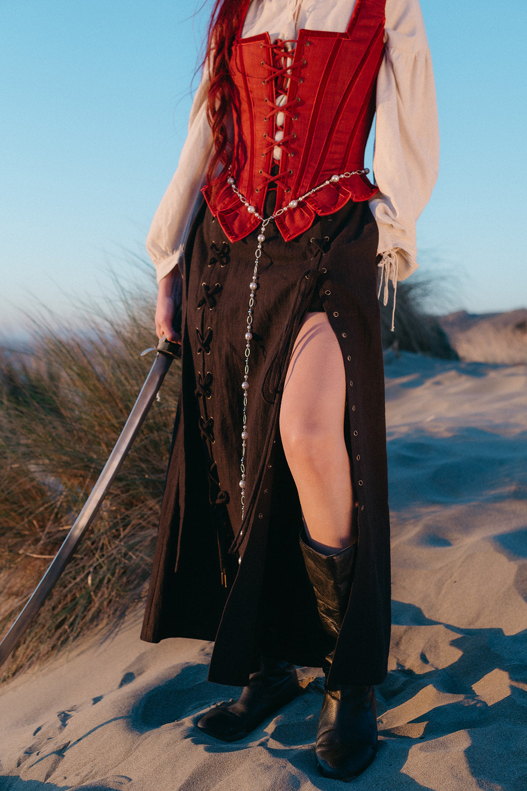 Woman wearing a black Tavern Skirt with side slit and leather boots standing on sandy dunes holding a sword