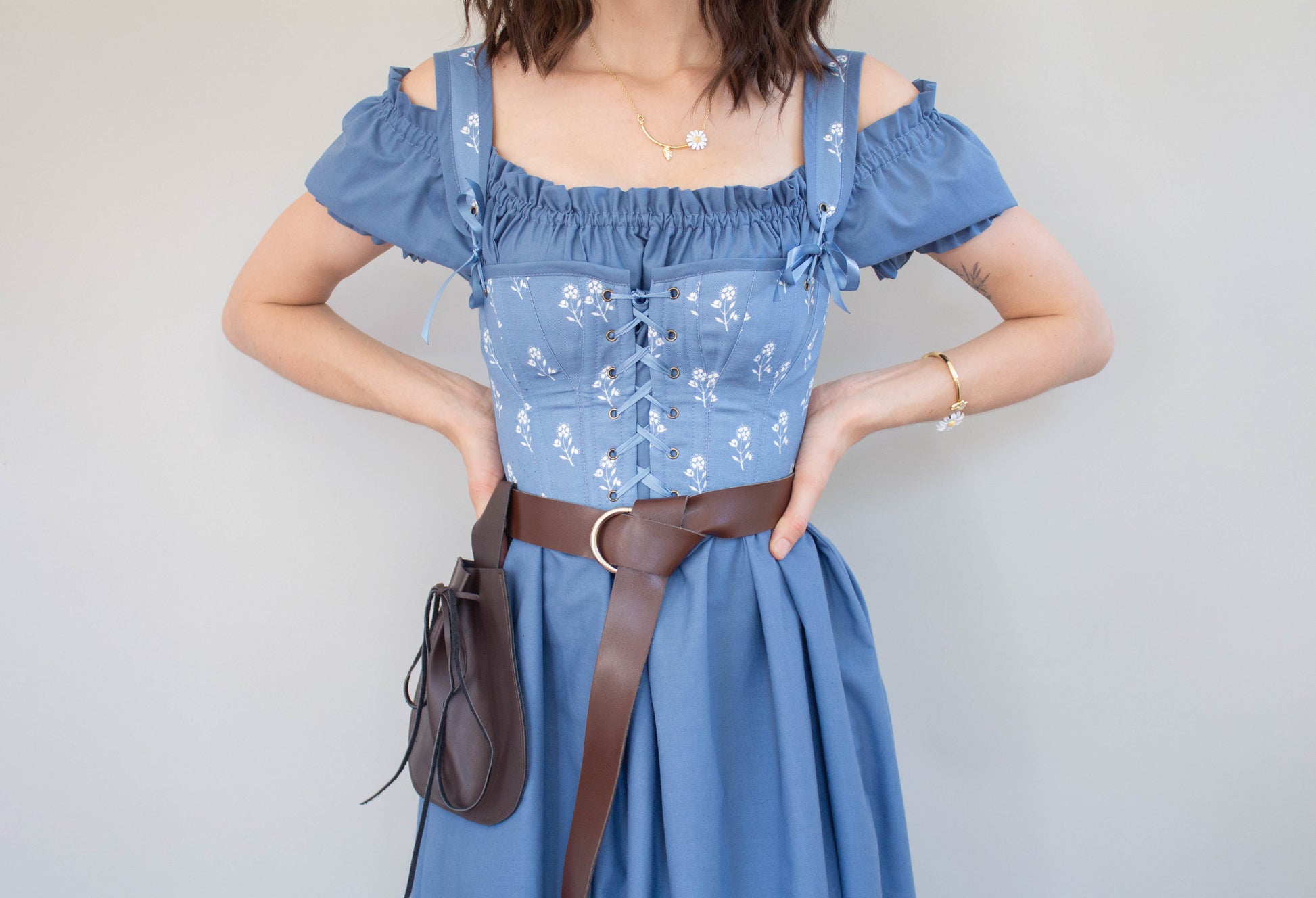 Woman wearing Handmade Blue and White Floral Regency Stays - Size XS with off-shoulder sleeves and a brown belt bag on gray background