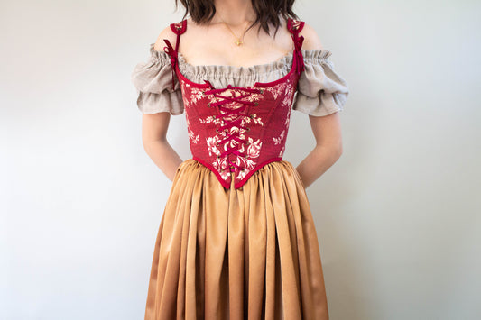 Woman wearing a Handmade Dark Red Toile Corset with floral patterns paired with a beige puff sleeve blouse and golden skirt