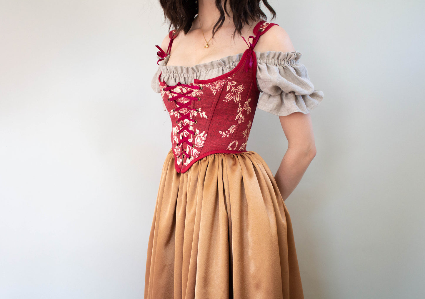Woman wearing a Handmade Dark Red Toile Corset over beige puffed sleeves with a flowing tan skirt on a white background