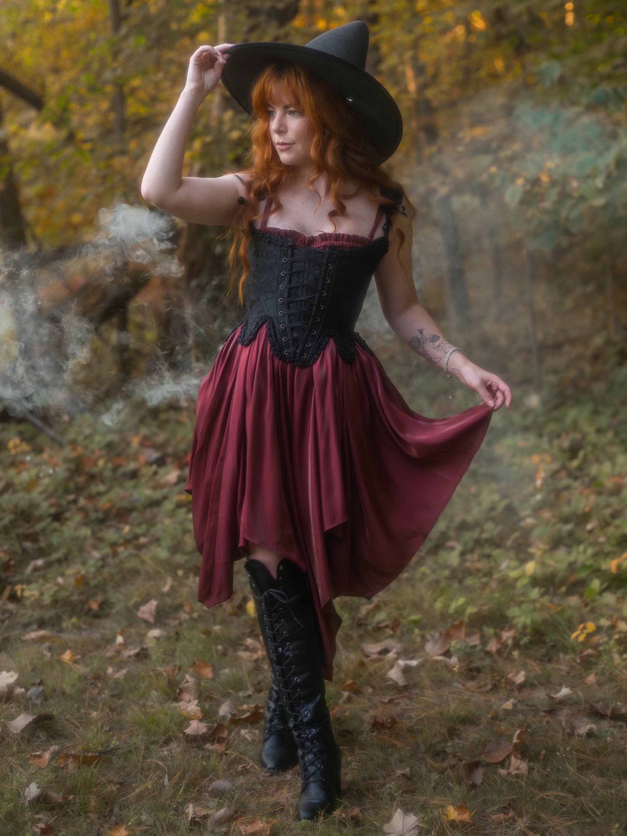 Woman with long red hair wearing a black witch hat, black corset, burgundy skirt, and black boots in a forest setting