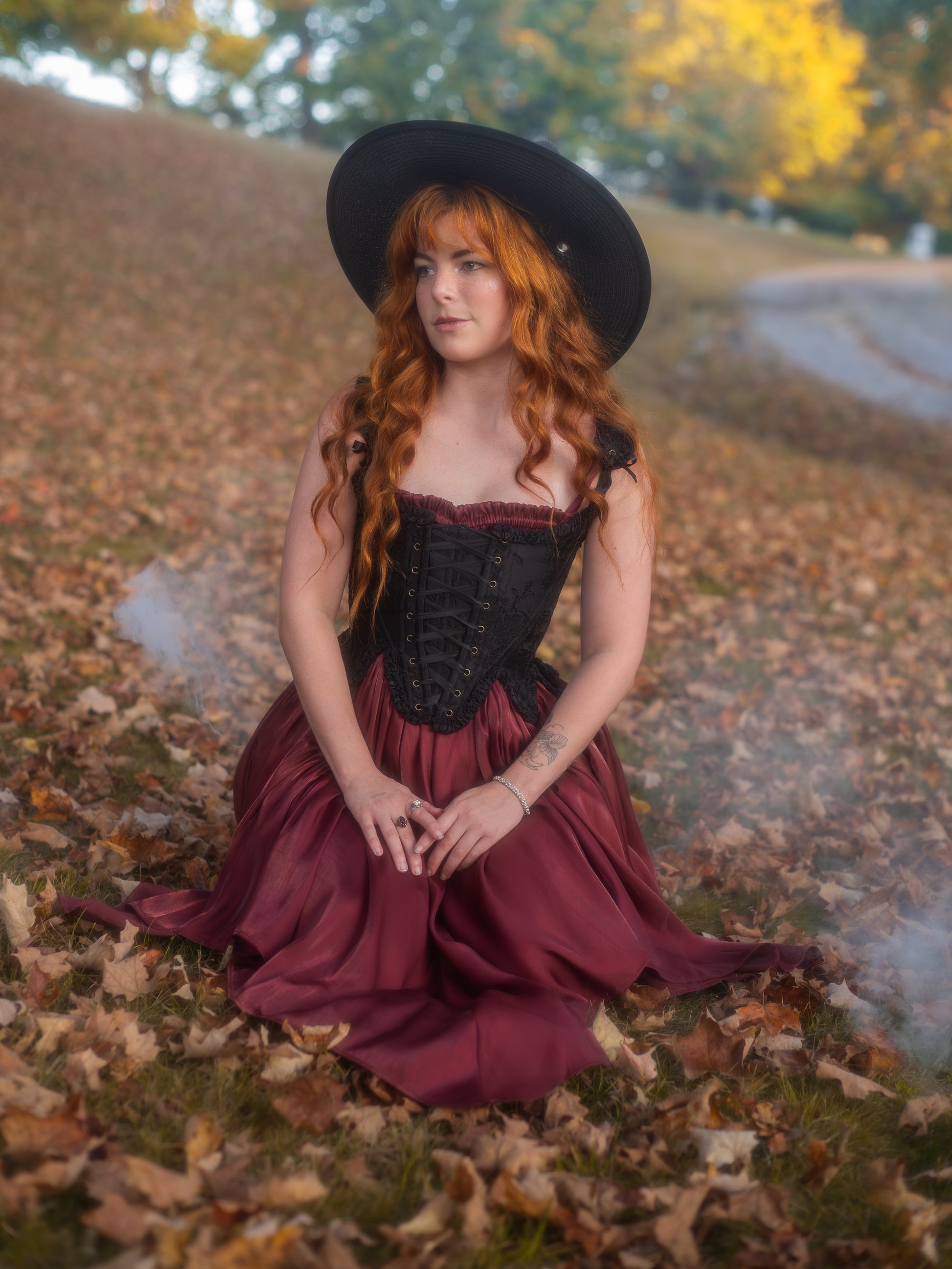 Woman with long red hair wearing a dark red flutter dress and black corset sitting among autumn leaves outdoors