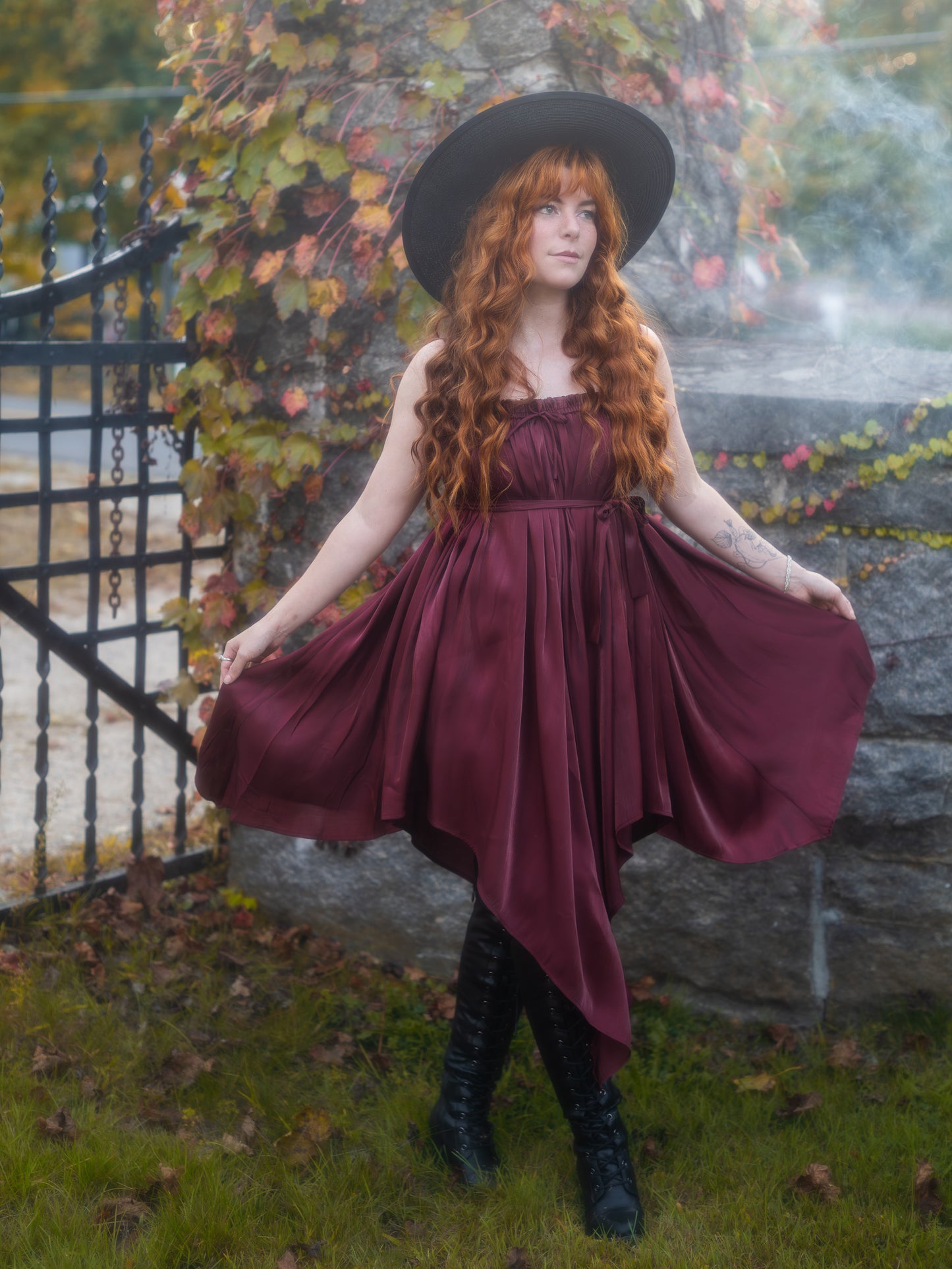 Woman with long red hair wearing a Dark Red Flutter Dress with handkerchief hem and black hat outdoors by a gate