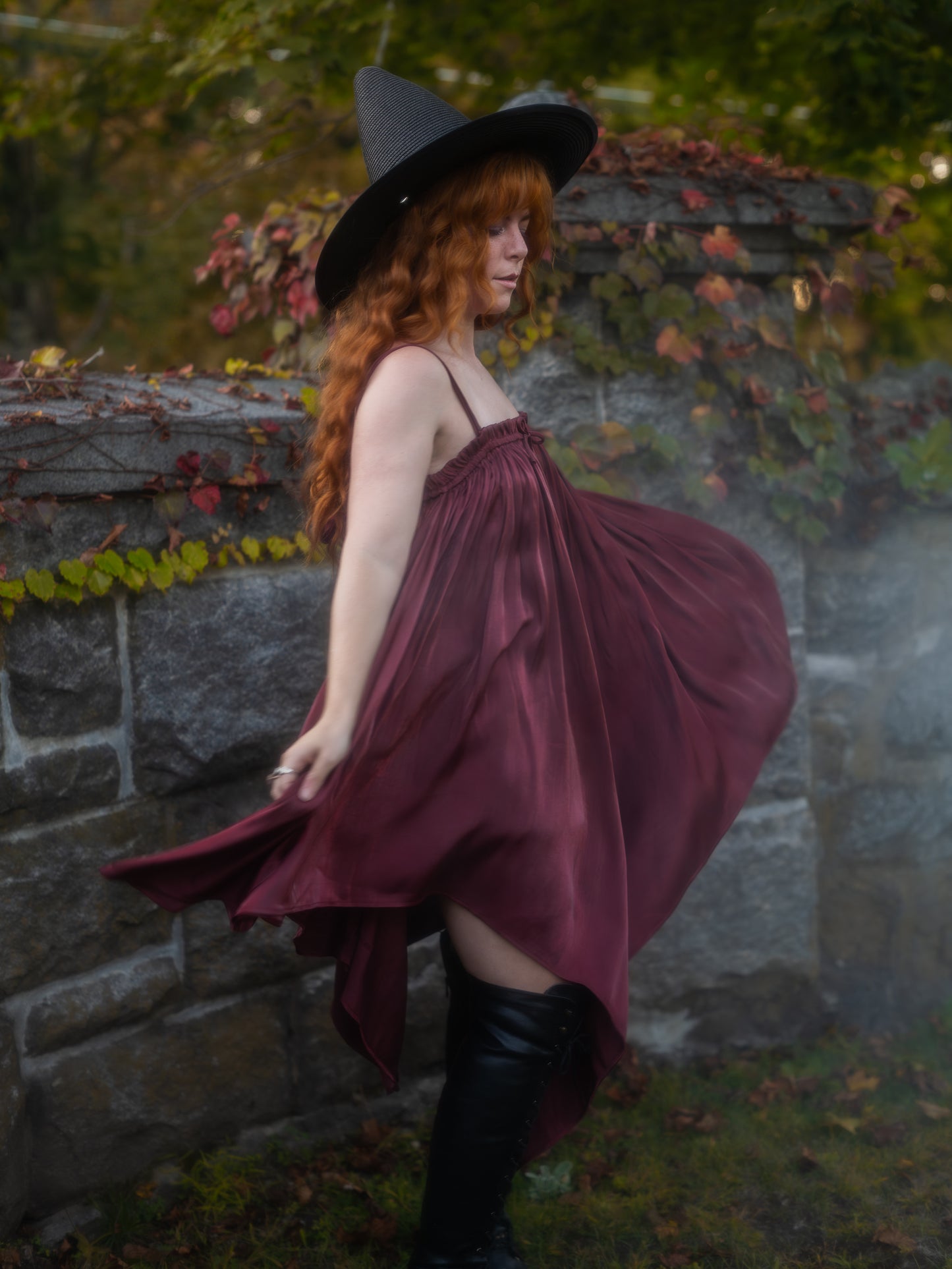 Woman wearing a Dark Red Flutter Dress with a handkerchief hem spinning outdoors near a stone wall with autumn leaves.