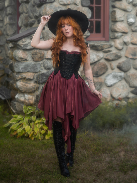 Woman wearing Dark Red Flutter Dress with handkerchief hem and black corset top styled with black boots and hat outdoors