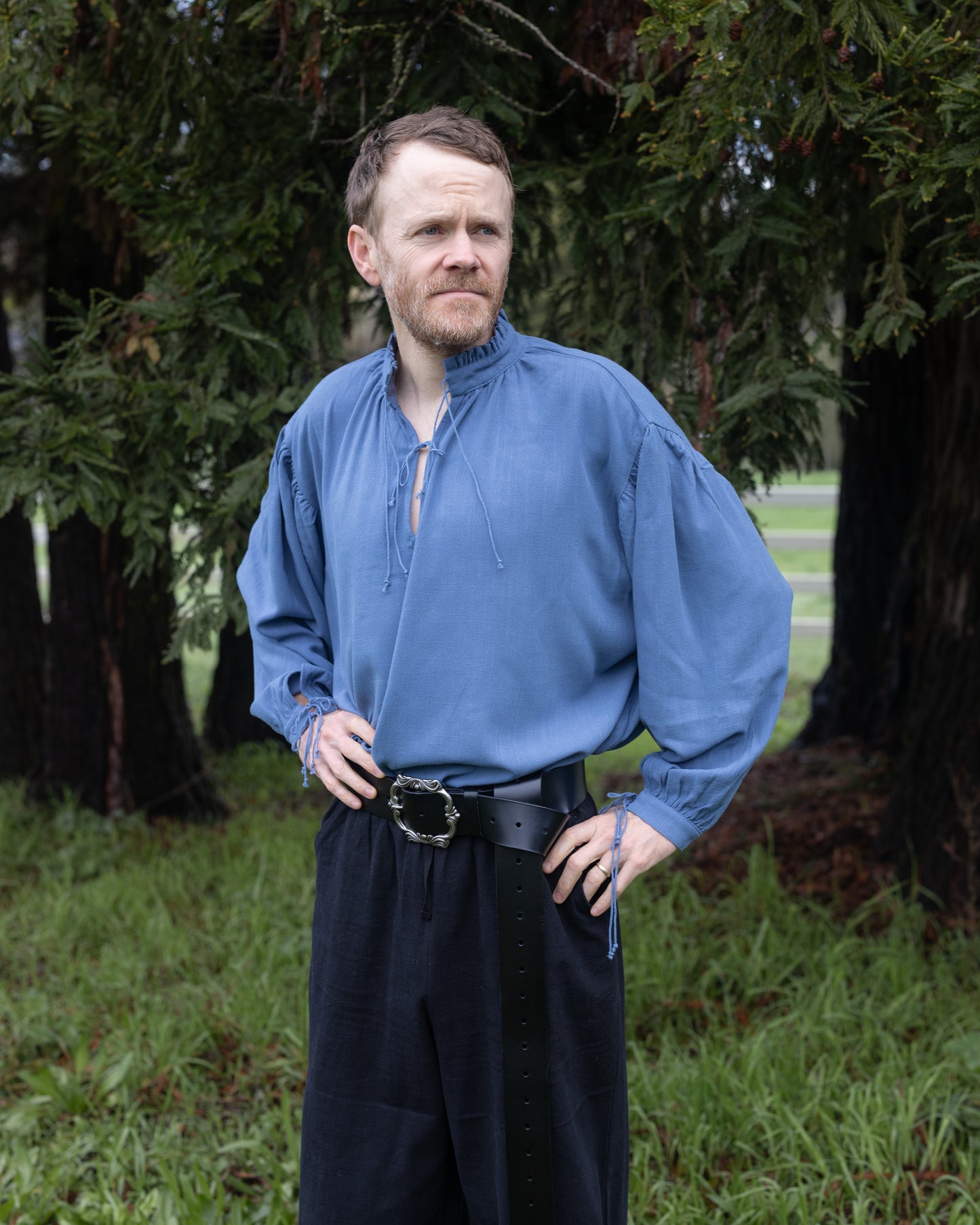Man wearing a blue Borgia Shirt with billowy sleeves and laced collar standing outdoors near trees