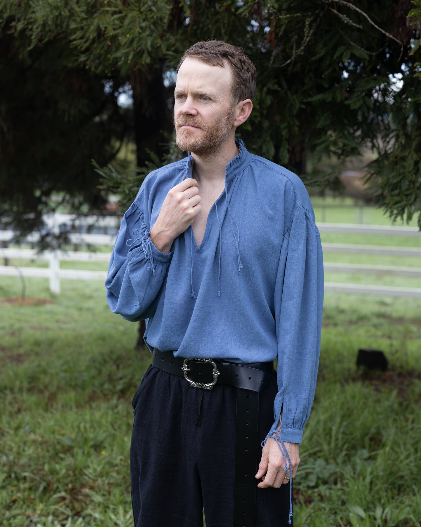 Man wearing blue Borgia Shirt with loose sleeves and drawstring neckline outdoors near trees and fence