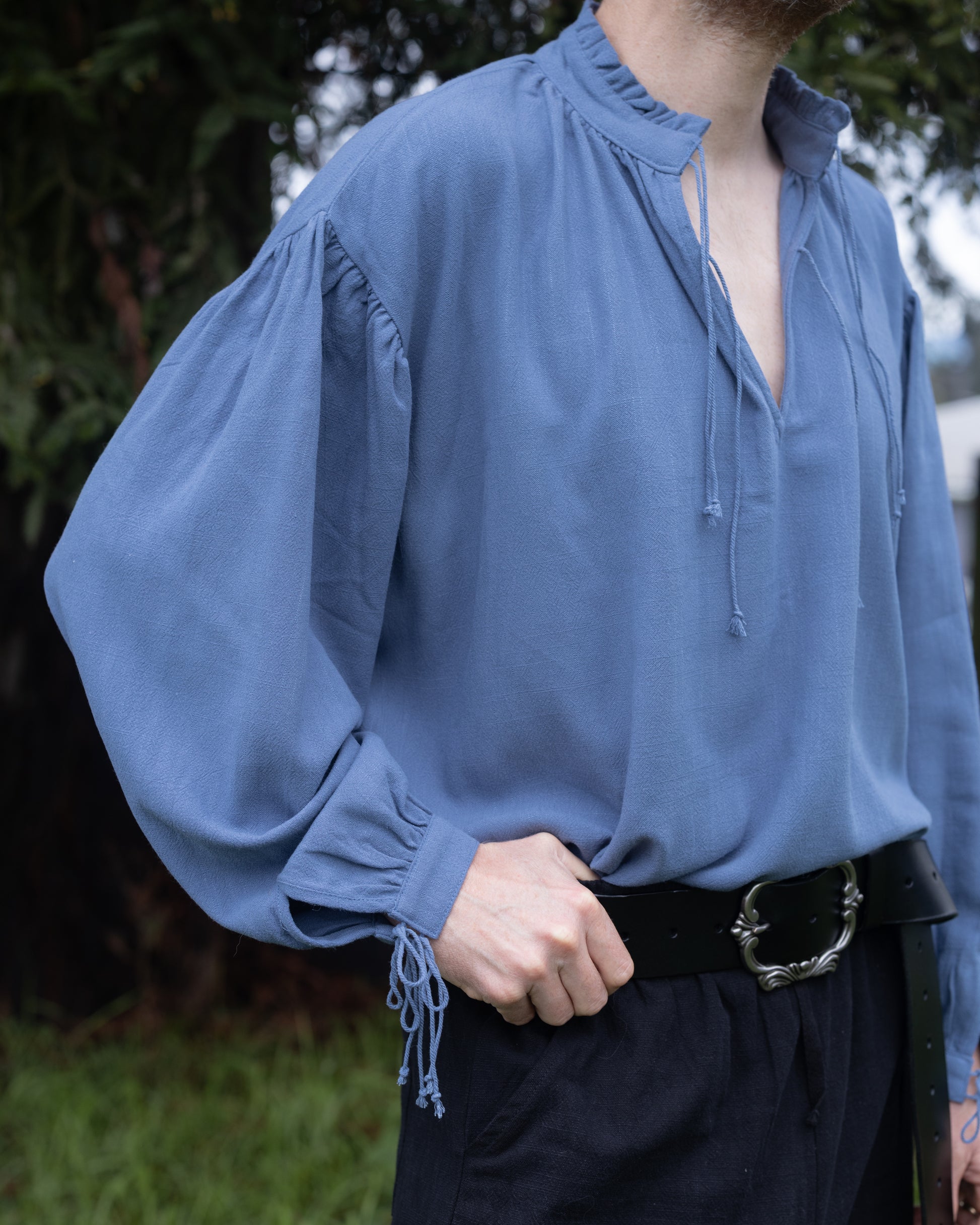 Man wearing a loose blue Borgia Shirt with billowy sleeves and a tied neckline in an outdoor setting