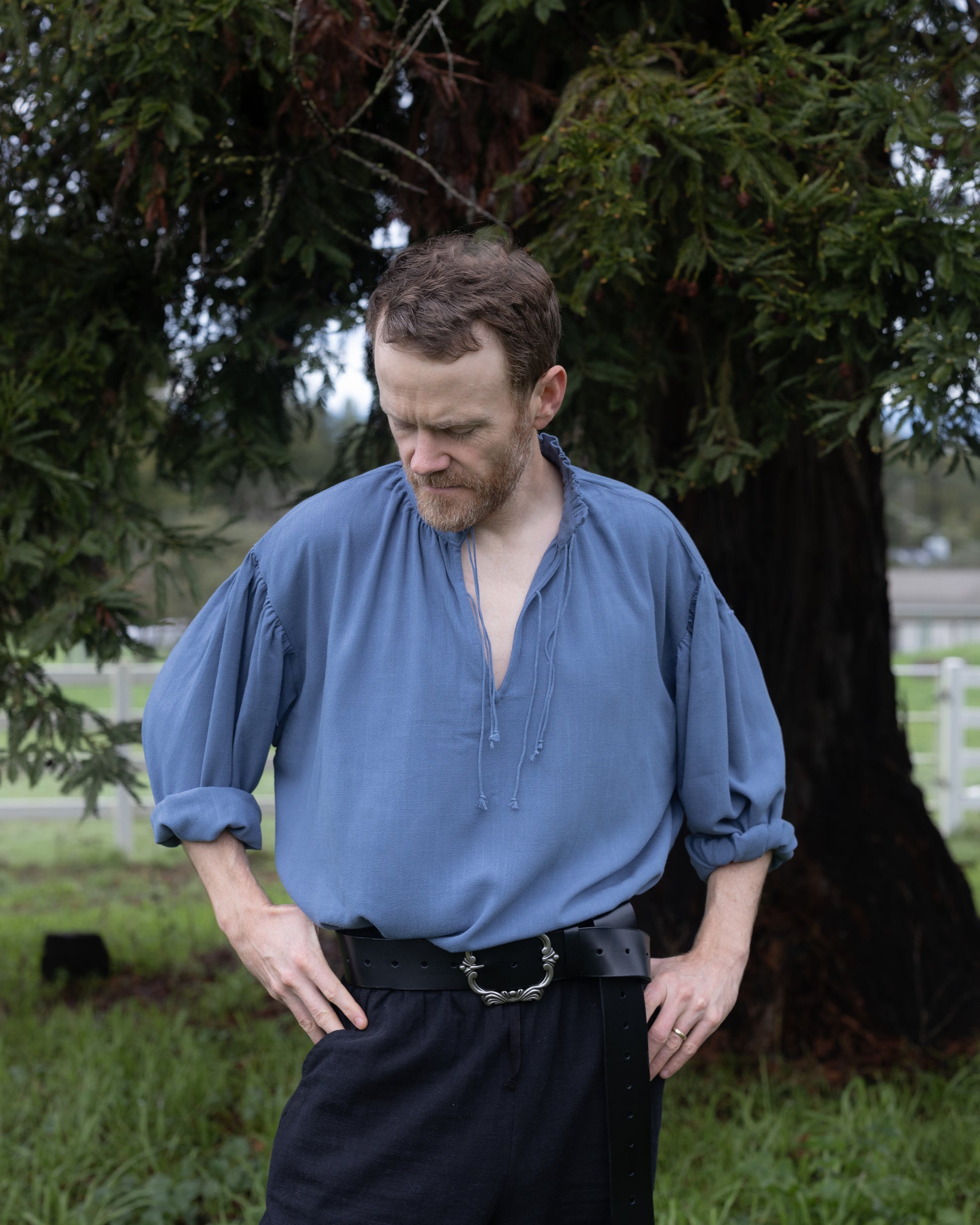 Man wearing blue Borgia Shirt with loose sleeves and open collar standing outdoors near tree
