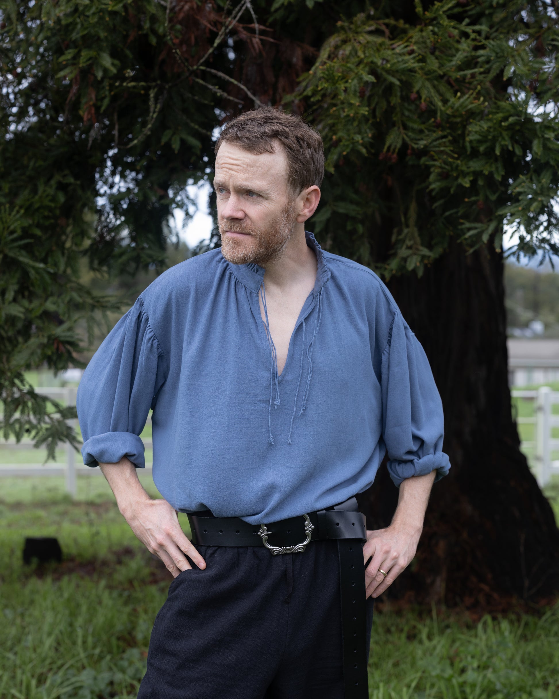 Man wearing a blue Borgia Shirt with rolled sleeves standing outdoors near a tree in a relaxed pose