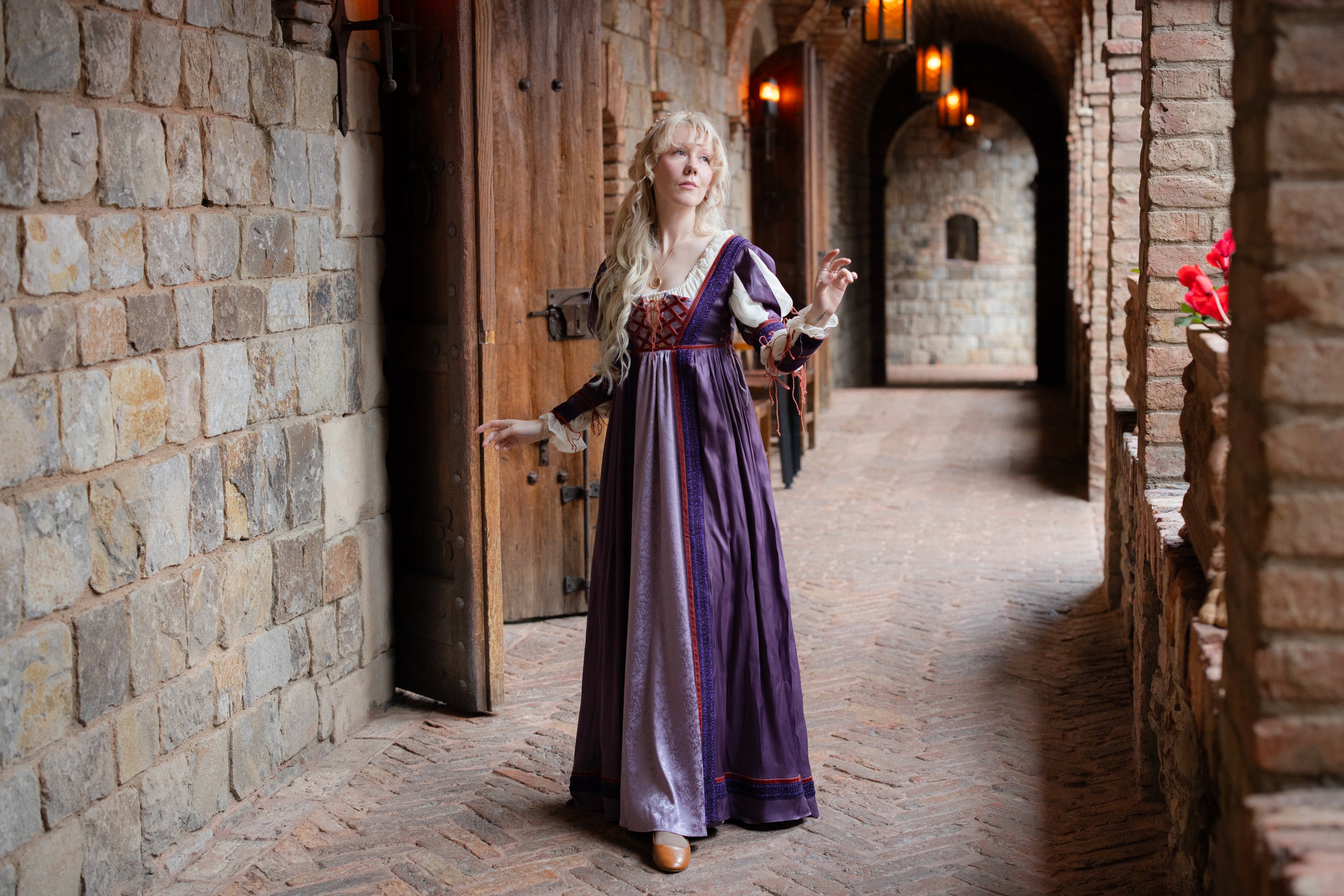 Blonde woman in a flowing purple and lavender Renaissance gown standing in a stone corridor with wooden doors