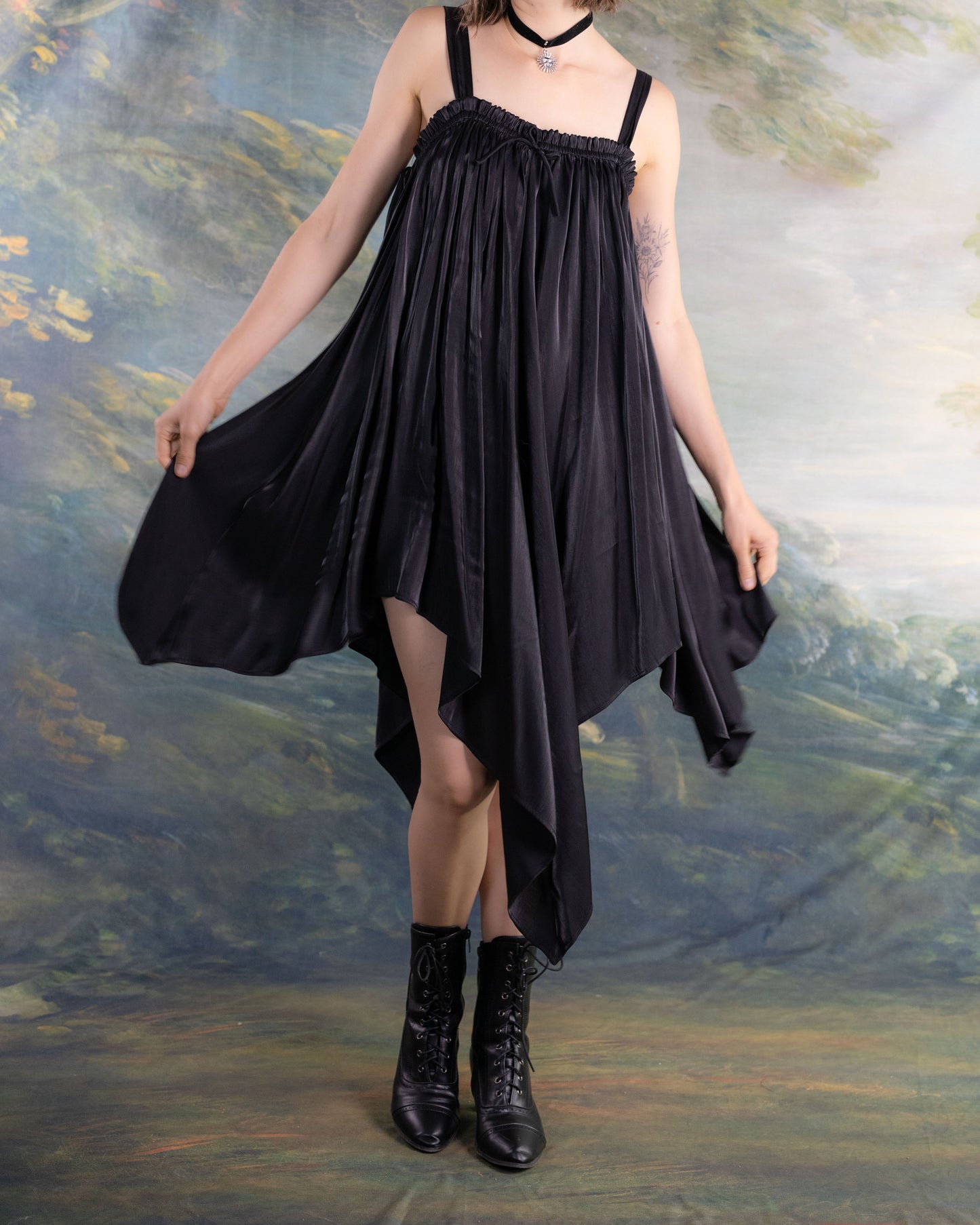 Woman wearing a flowing Black Flutter Fairy Dress with handkerchief hem and black boots against a soft nature backdrop