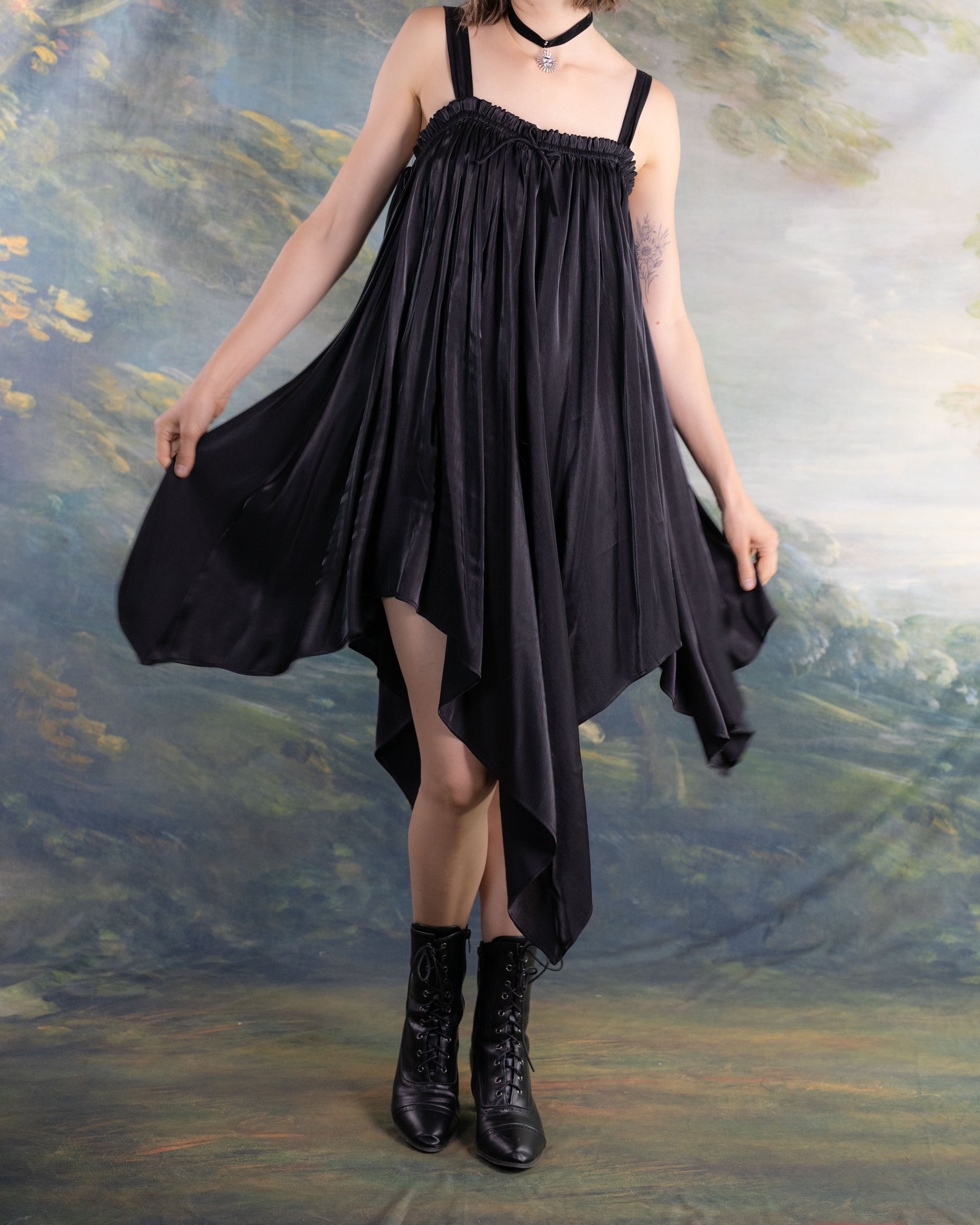 Woman wearing a flowing Black Flutter Fairy Dress with handkerchief hem and black boots against a soft nature backdrop