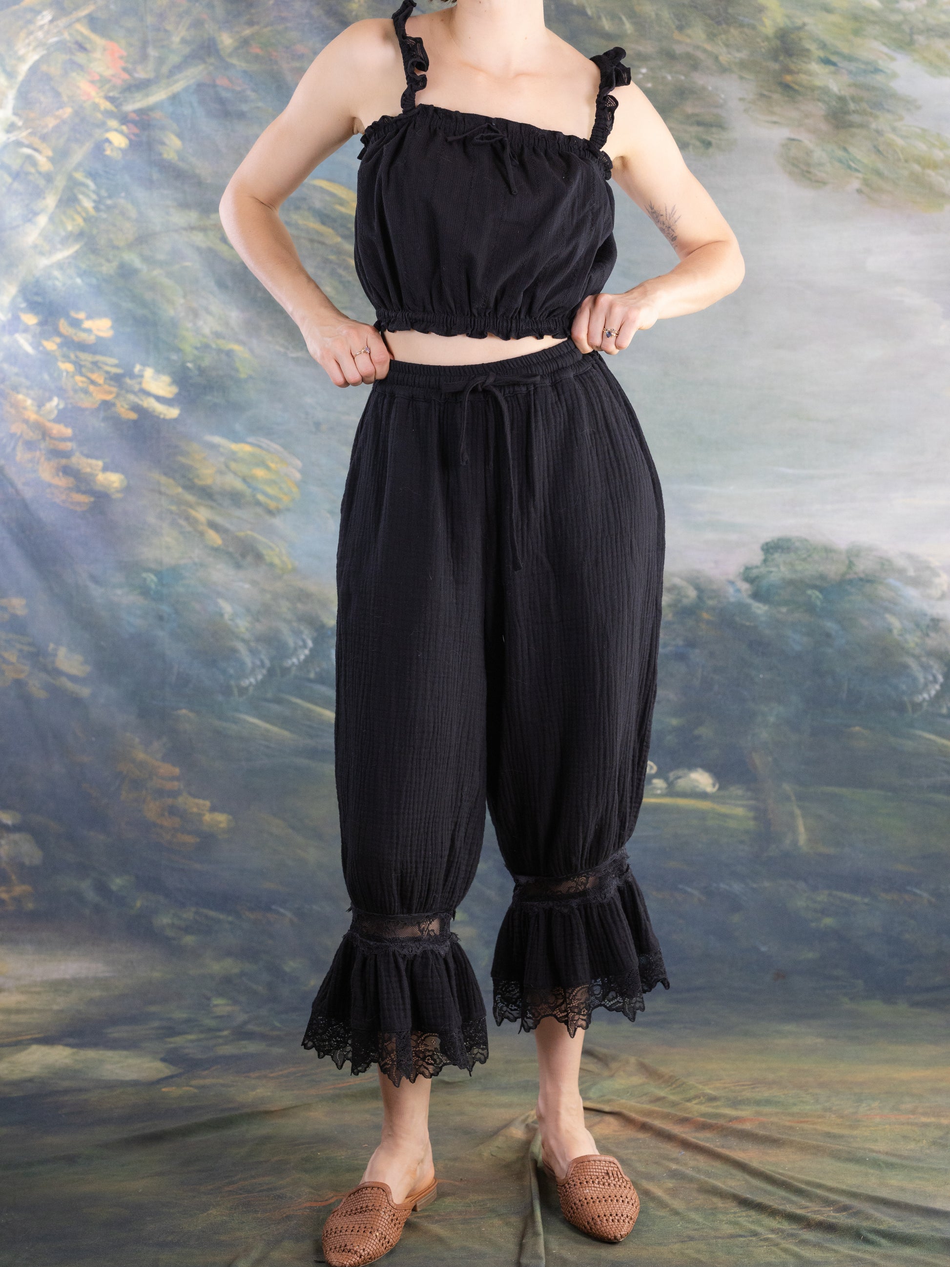 Model wearing black Lacey Chemisette Tank with textured pants featuring lace details at the hem against artistic backdrop
