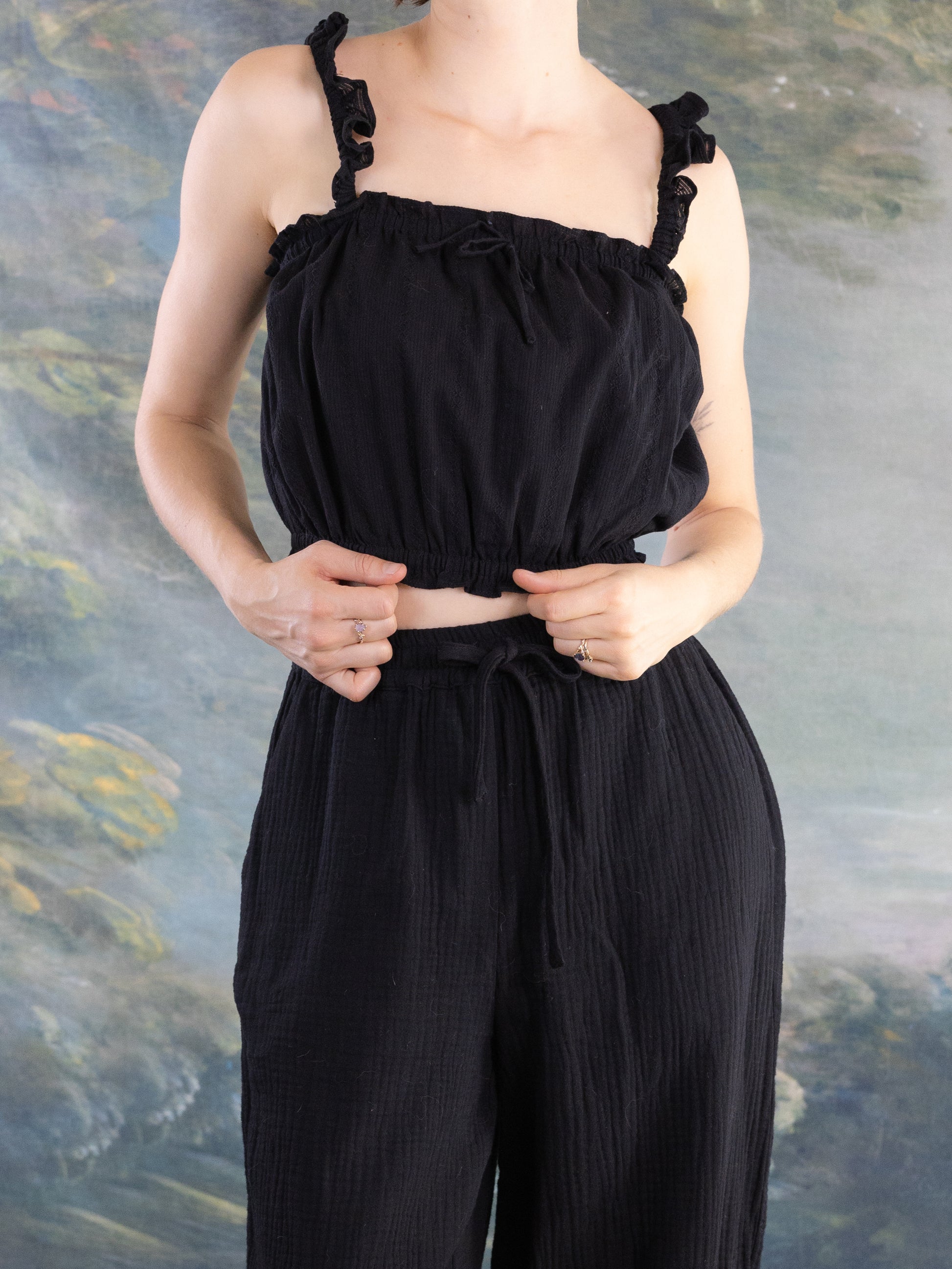 Woman wearing black Lacey Chemisette Tank with ruffled straps and matching high-waisted pants against painted backdrop