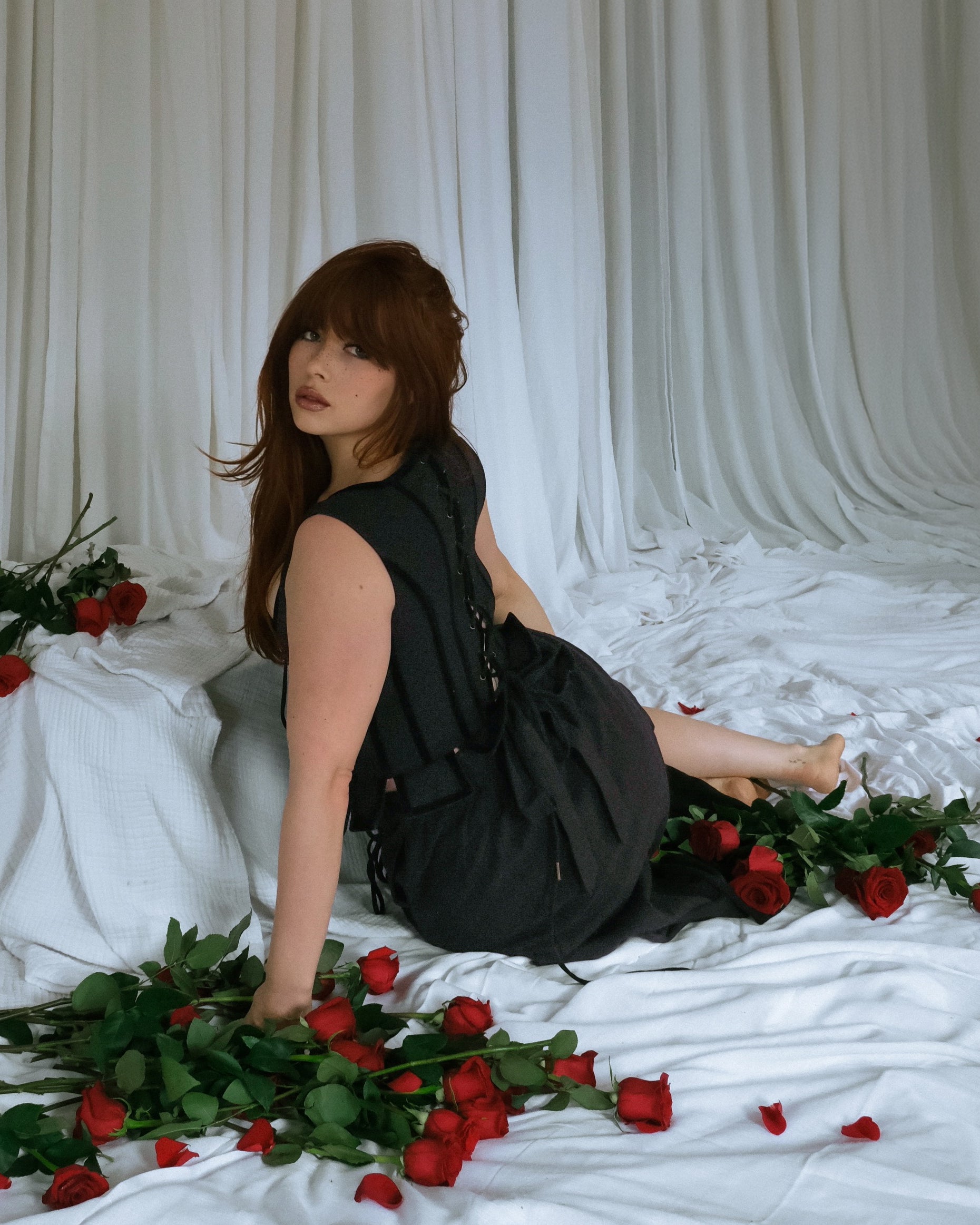 Woman wearing Black Pirate Doublet sitting on white fabric surrounded by scattered red roses and green leaves