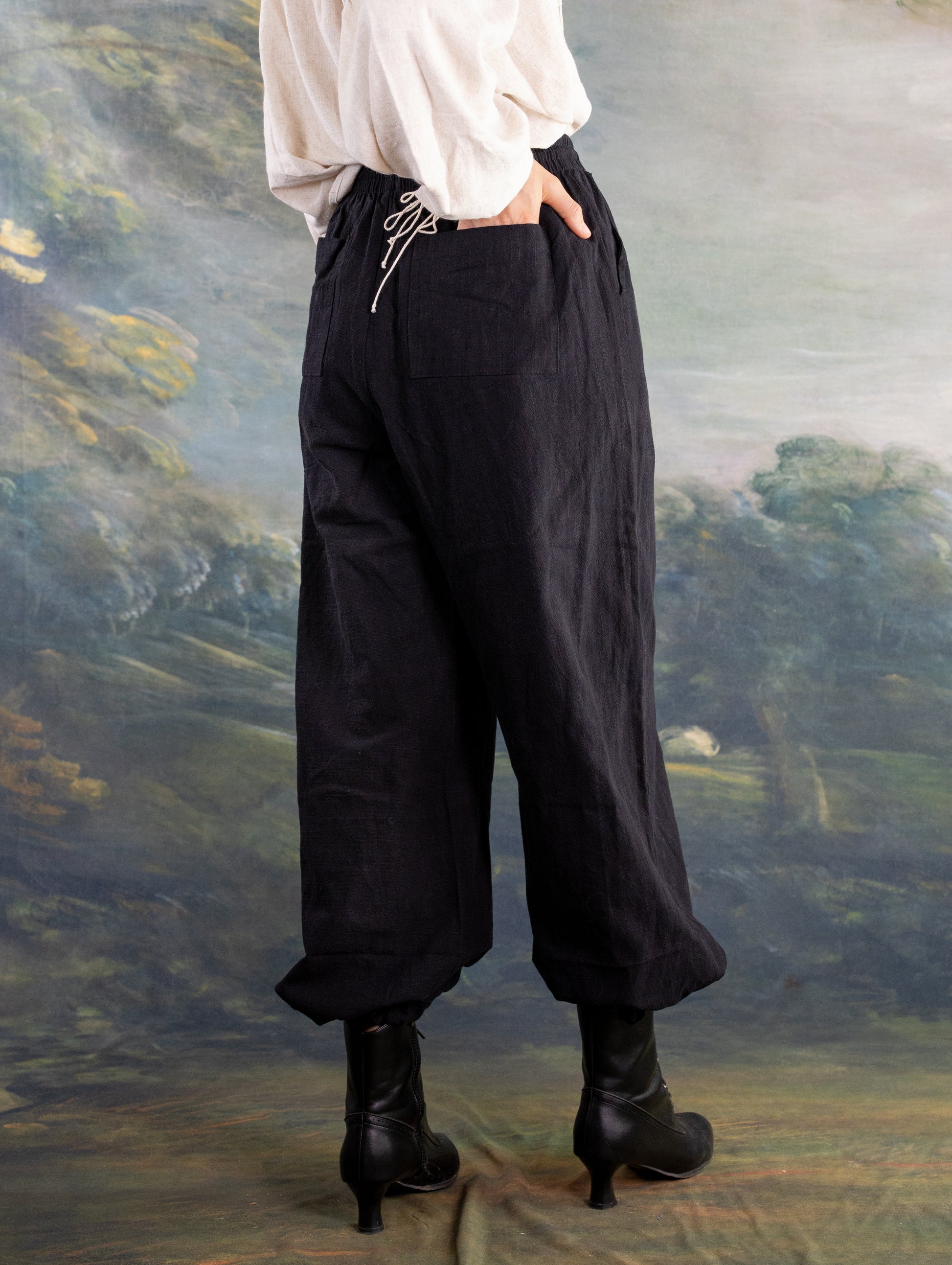 Back view of rustic black Pirate Pants worn with heeled boots and a loose white top against a painted backdrop
