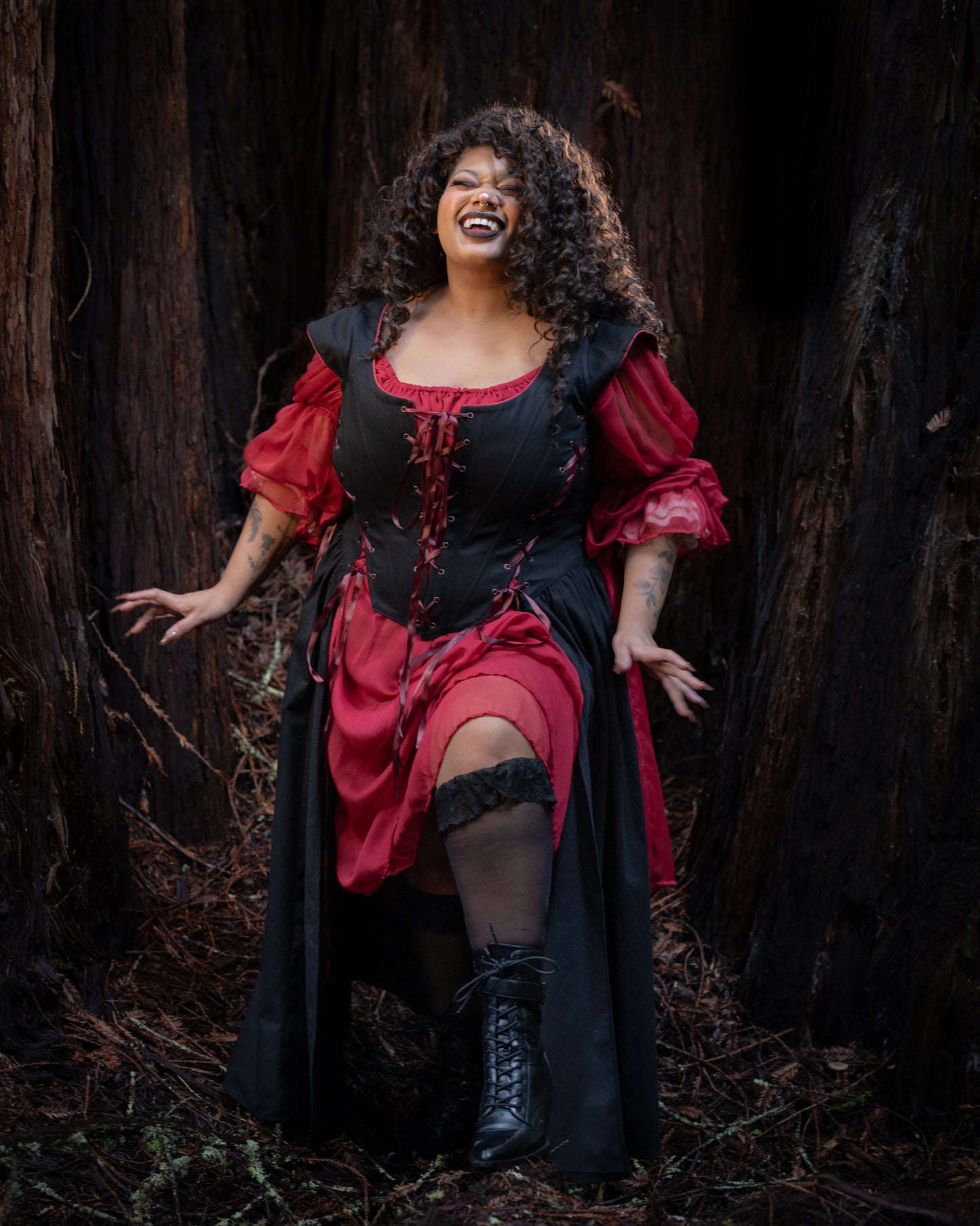 Woman in a Black Vampire Slayer Corset Dress with red sleeves and black boots standing in a dark forest setting smiling