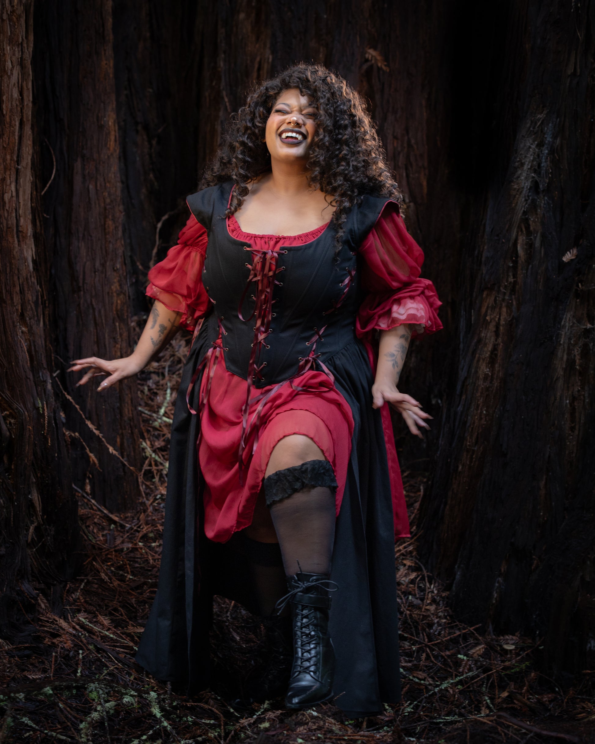 Woman in a Black Vampire Slayer Corset Dress with red sleeves and black boots standing in a dark forest setting smiling