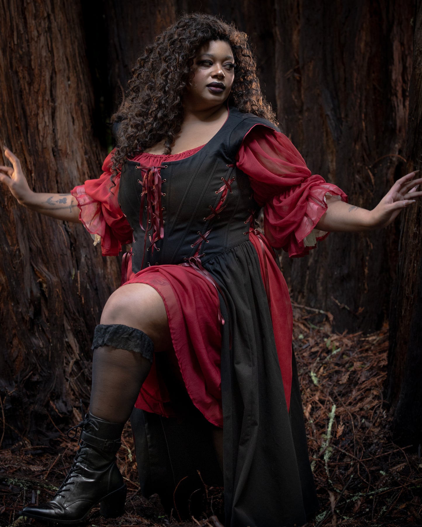 Woman wearing Black Vampire Slayer Corset Dress with red sleeves posing confidently outdoors by a large tree trunk