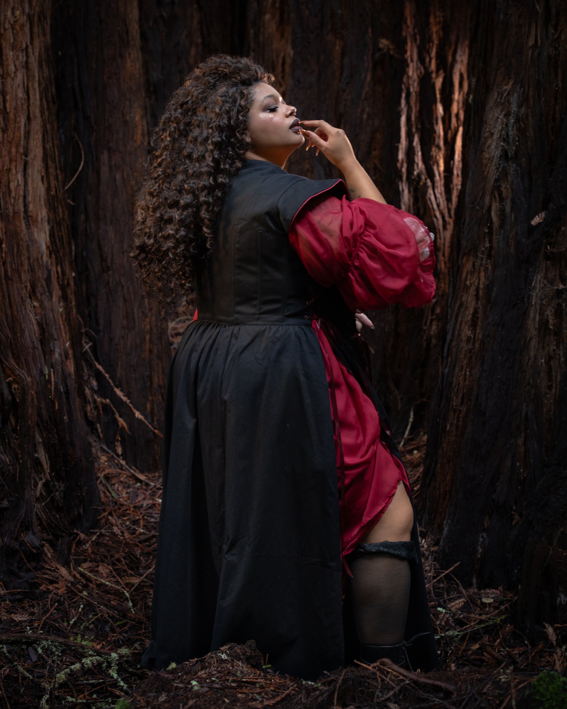 Woman with curly hair wearing Black Vampire Slayer Corset Dress standing thoughtfully among tall trees in forest setting