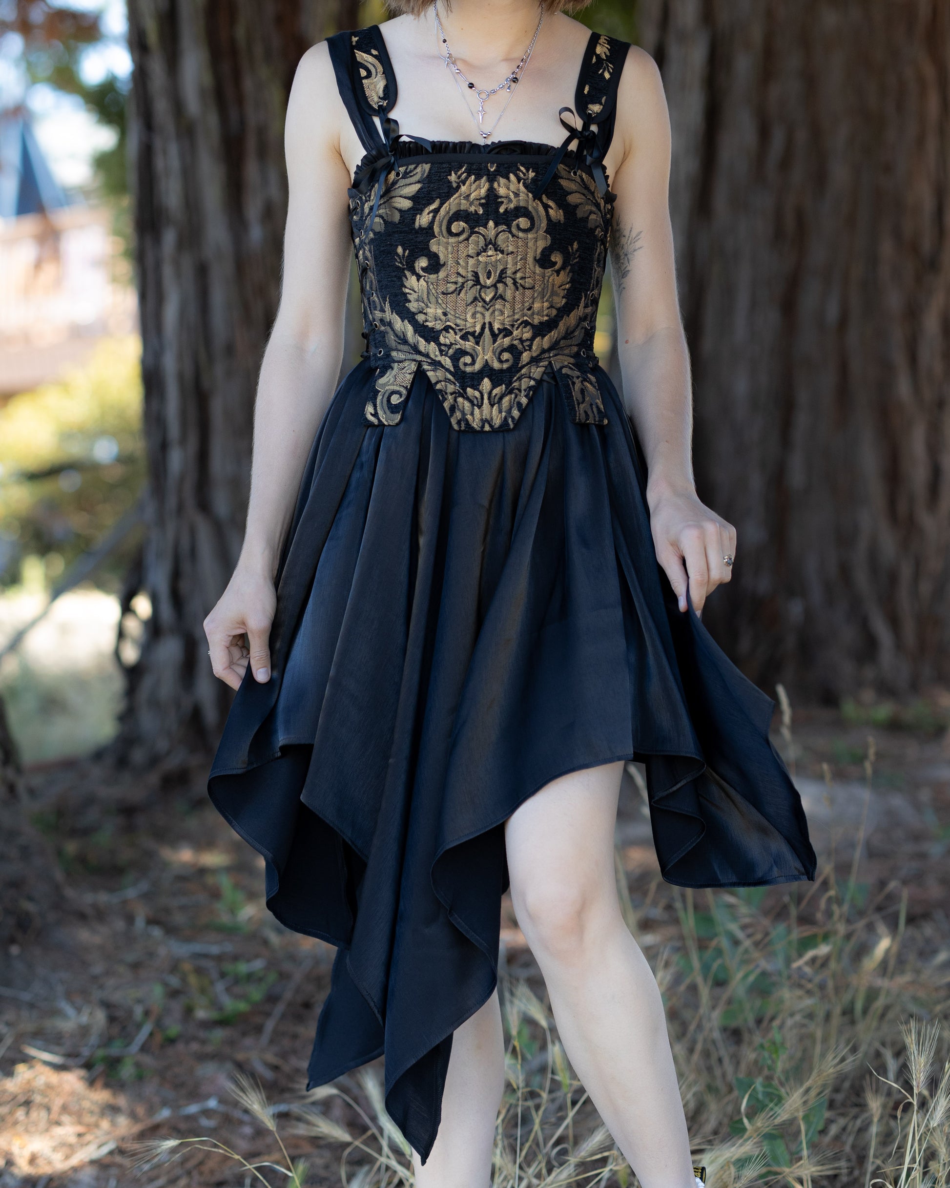Black Flutter Fairy Dress with ornate gold bodice and flowing black handkerchief hem in natural outdoor setting