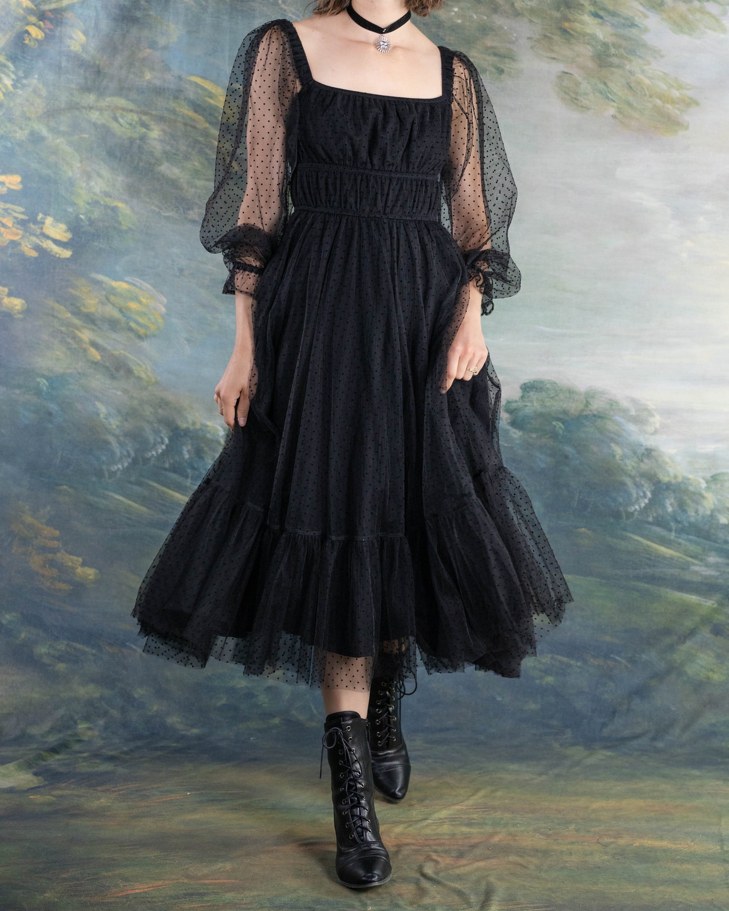 Woman wearing a Black Tulle Dot Dress with sheer puff sleeves and lace-up black boots against a painted backdrop