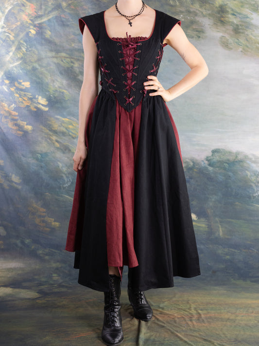 Woman wearing Black Vampire Slayer Corset Dress with lace-up bodice over maroon dress and black boots in studio setting