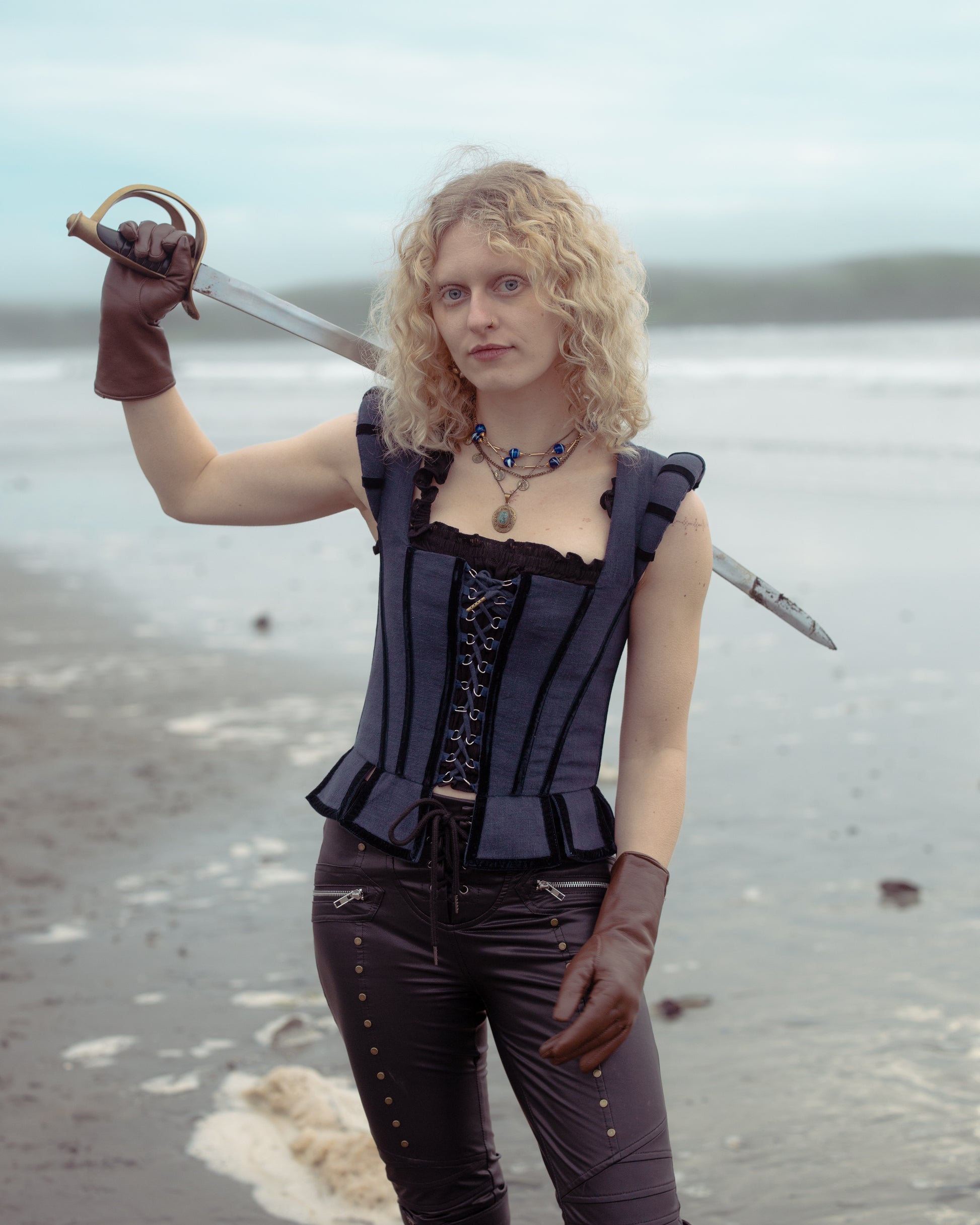 Woman wearing Navy Tavern Bodice holding sword over shoulder on beach with cloudy sky background