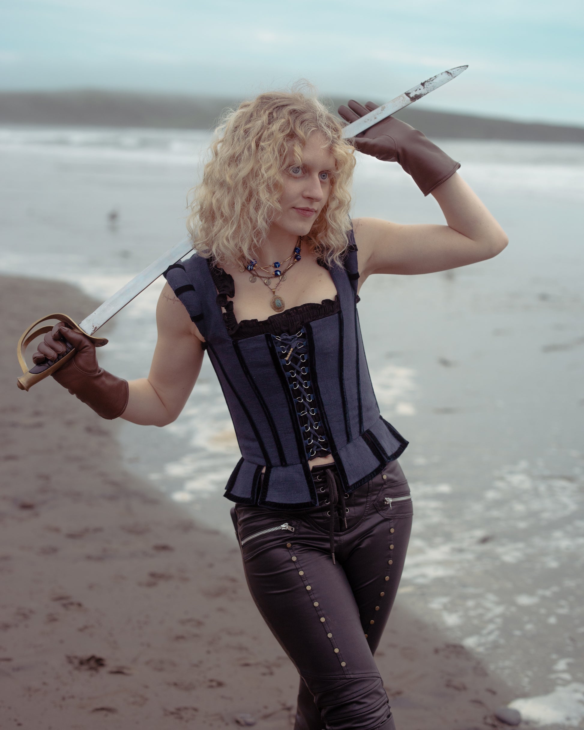 Woman wearing Navy Tavern Bodice and leather gloves posing with sword on a beach with waves in the background
