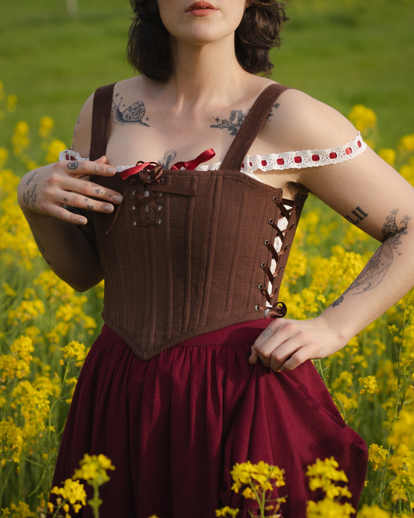 Close-up of a person wearing Brown Aurora Stays corset with lace-up sides and red skirt in a yellow flower field