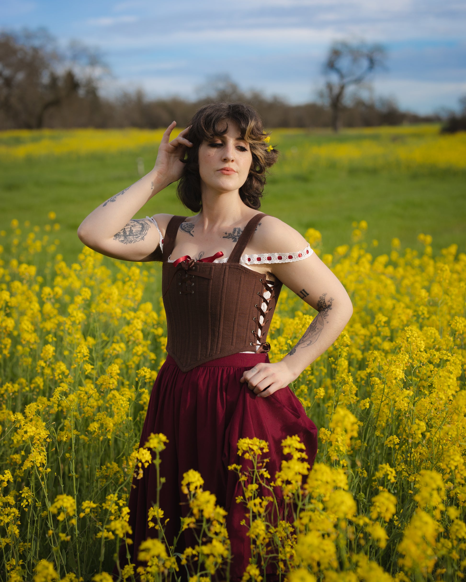 Woman wearing a Brown Aurora Stays corset with a burgundy skirt standing in a field of yellow flowers under blue sky