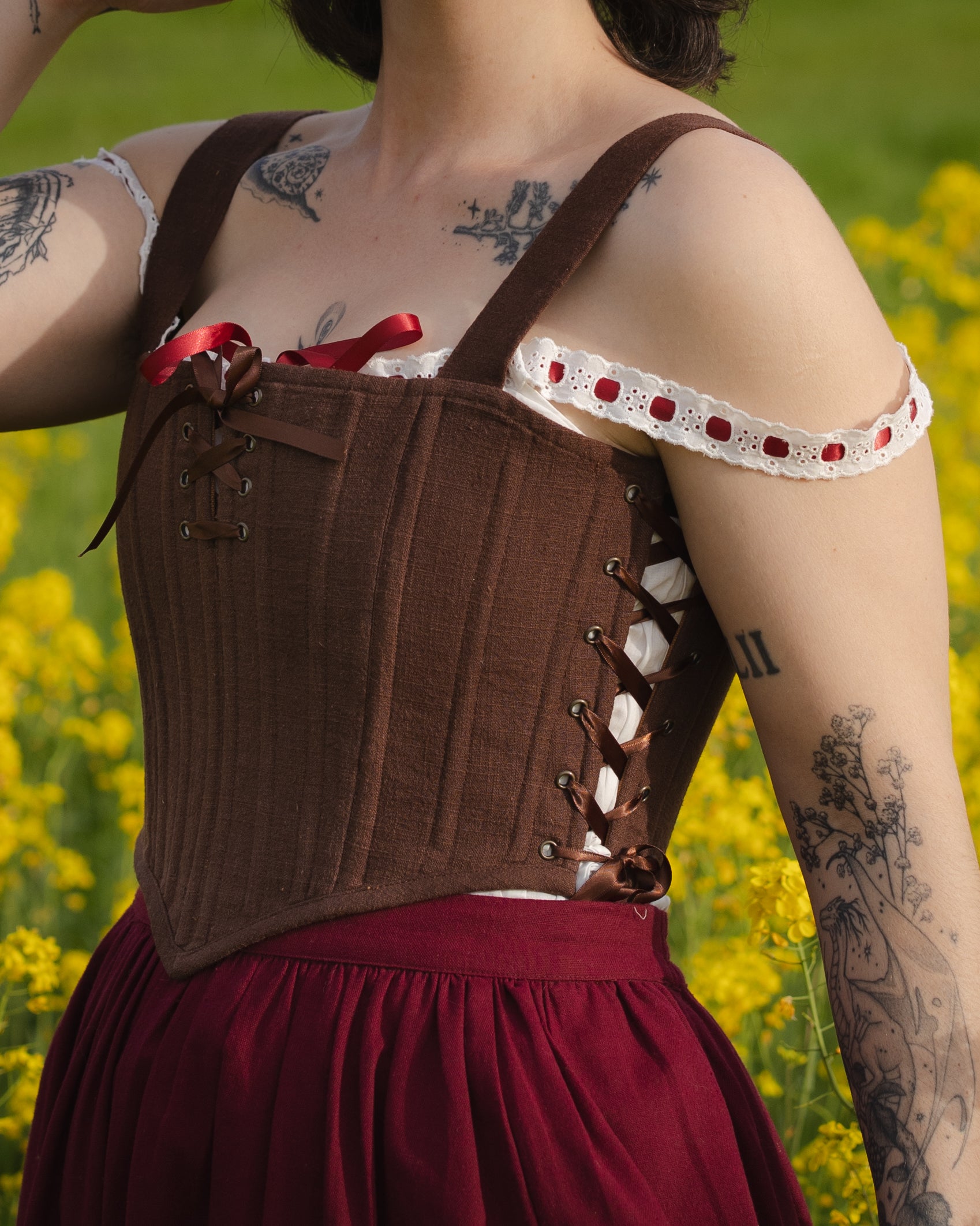 Close-up of a person wearing brown corset with lace-up sides and red ribbon details called Brown Aurora Stays outdoors