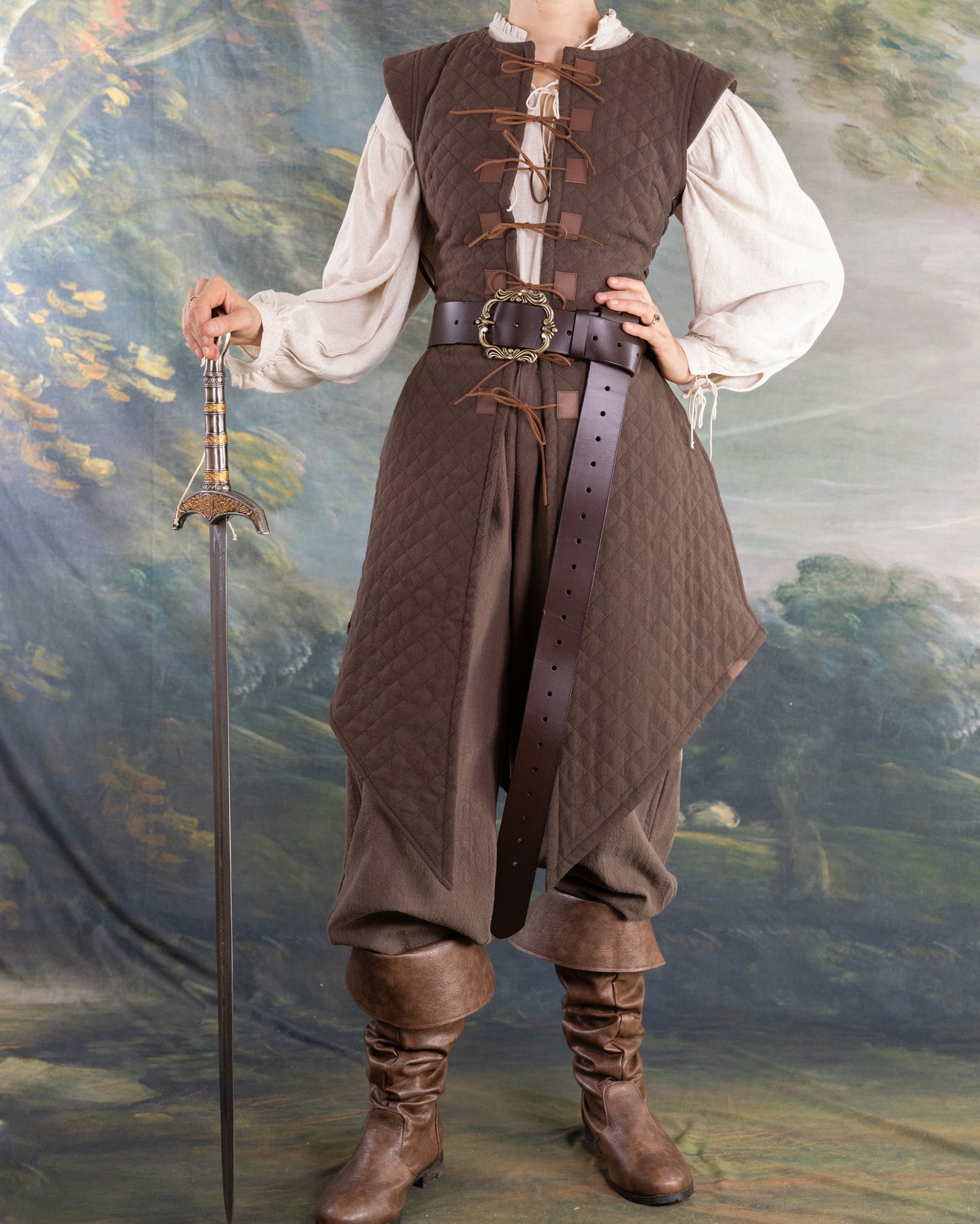 Woman wearing a brown Quilted Gambeson with leather belt and boots holding a medieval-style sword against a painted backdrop