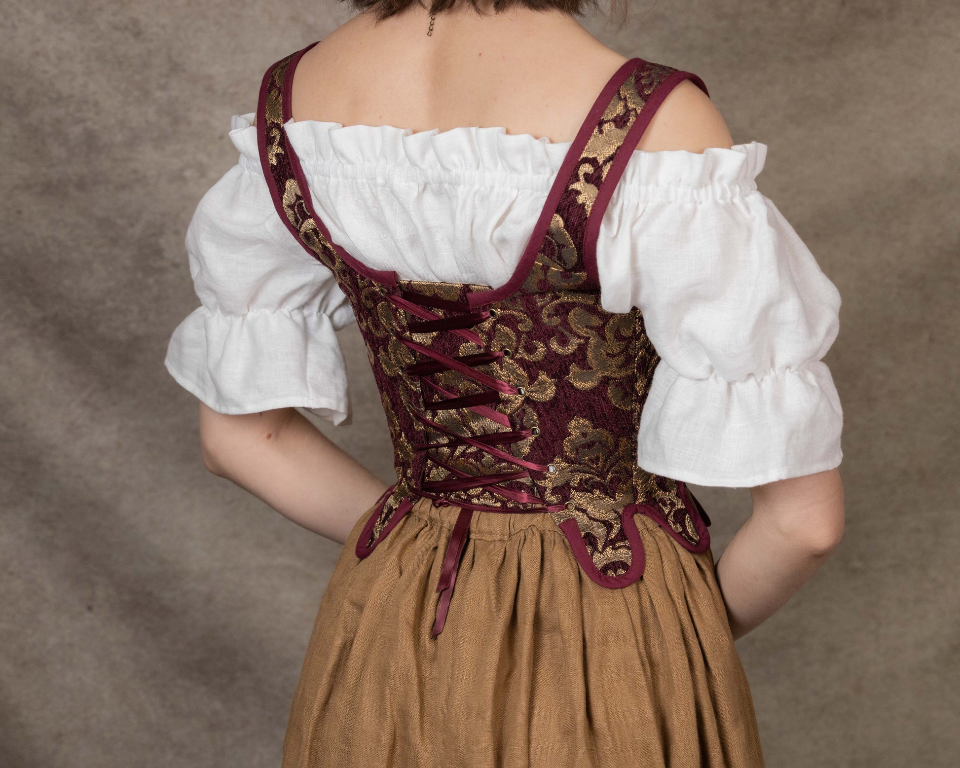 Back view of a woman wearing Handmade Burgundy Chenille Tudor Stays with gold floral patterns and a white blouse.