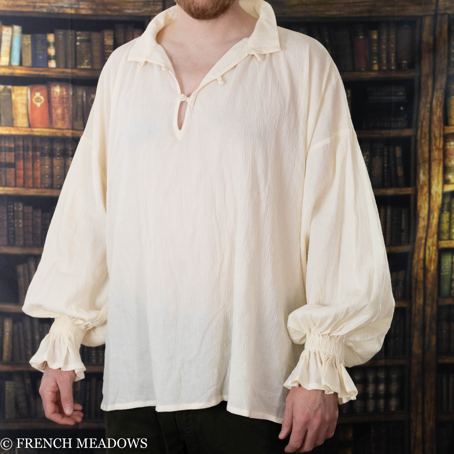Man wearing loose cream-colored Renaissance-style shirt with gathered cuffs standing in front of wood bookshelf filled with old books