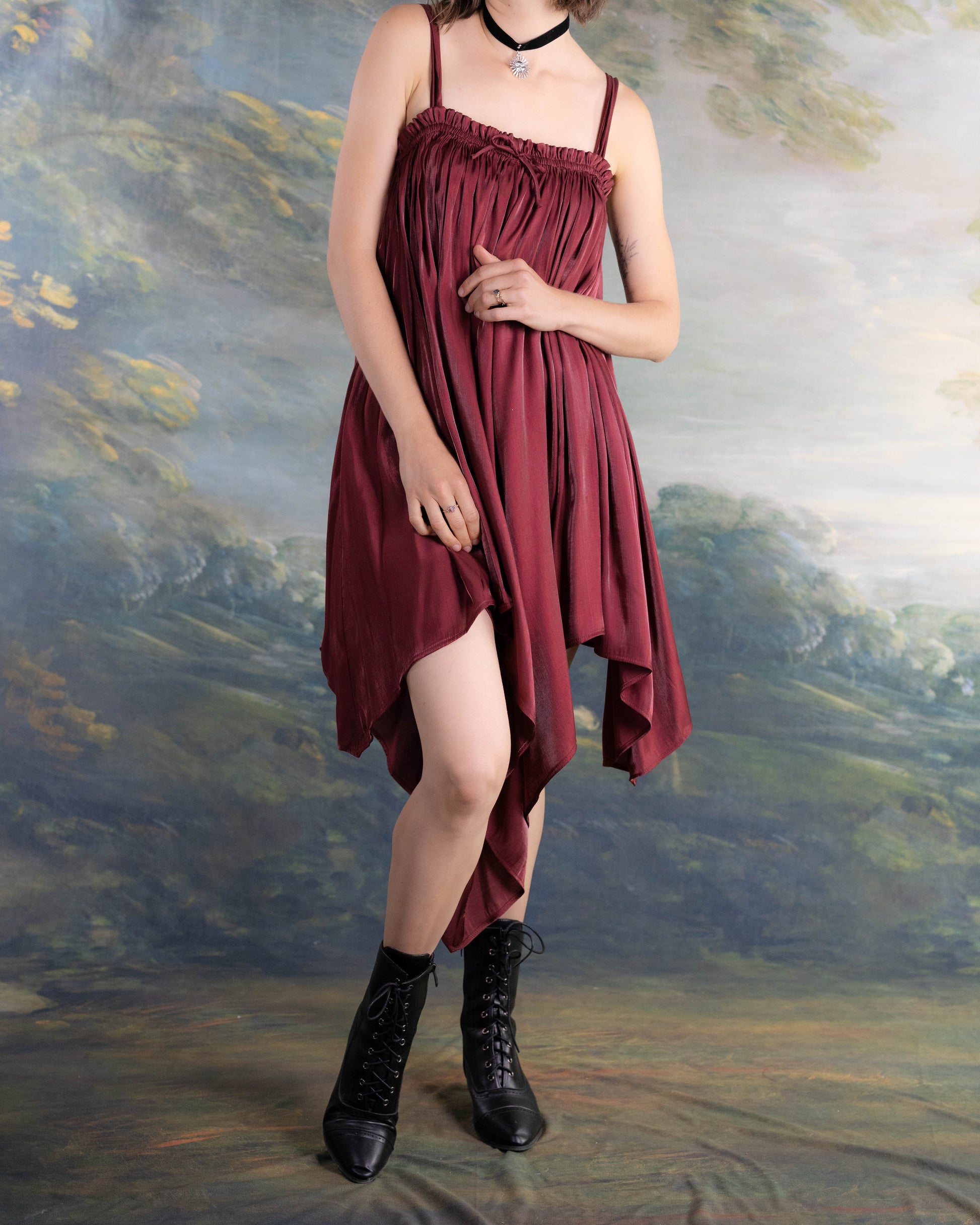 Woman wearing a Dark Red Flutter Dress with handkerchief hem and black lace-up boots against a painted backdrop