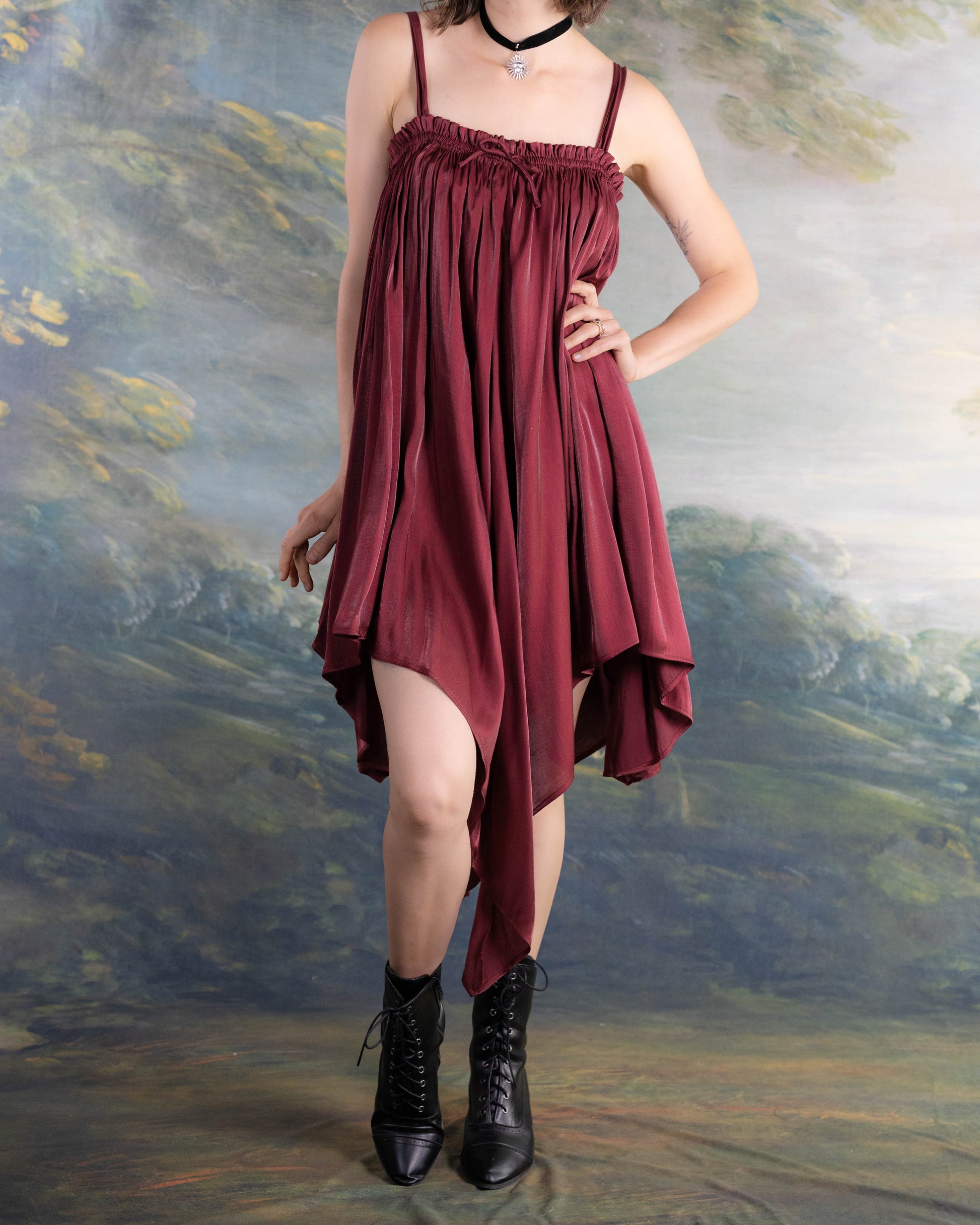 Woman wearing a Dark Red Flutter Dress with a handkerchief hem and black ankle boots, standing against a painted backdrop