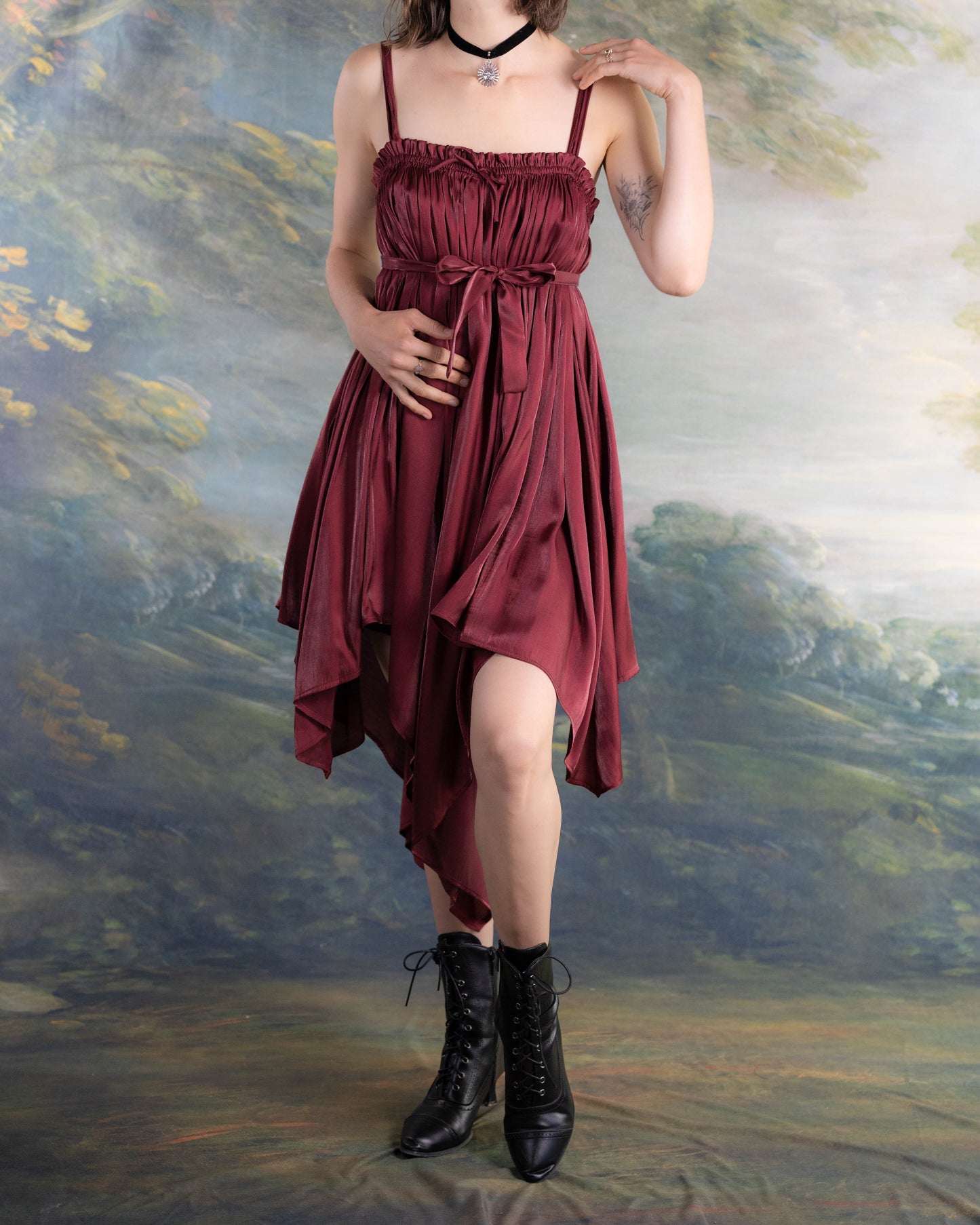 Woman wearing a dark red satin Dark Red Flutter Dress with handkerchief hem and black lace-up boots standing outdoors