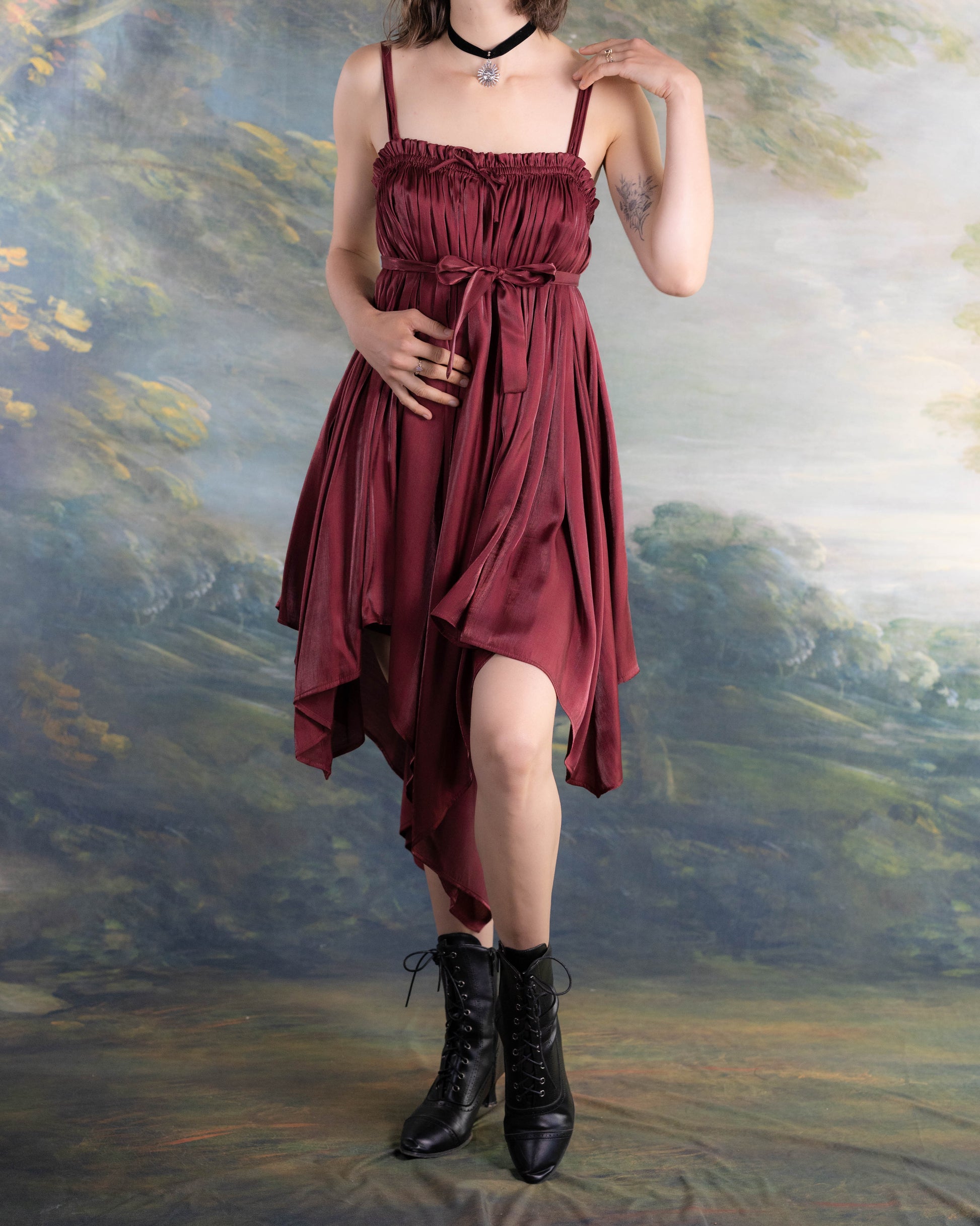 Woman wearing a dark red satin Dark Red Flutter Dress with handkerchief hem and black lace-up boots standing outdoors