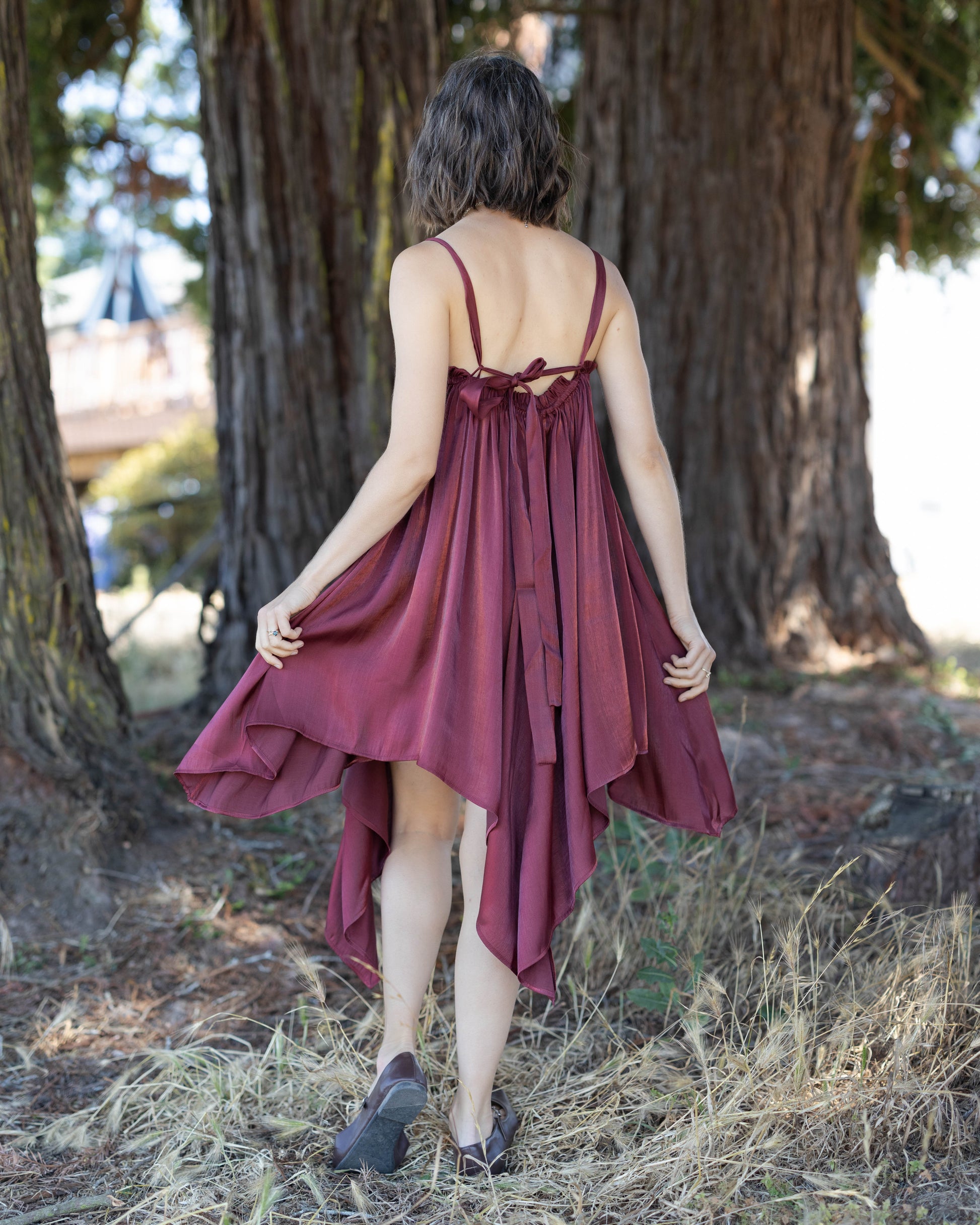 Woman wearing dark red flutter dress with handkerchief hem standing outdoors by large trees