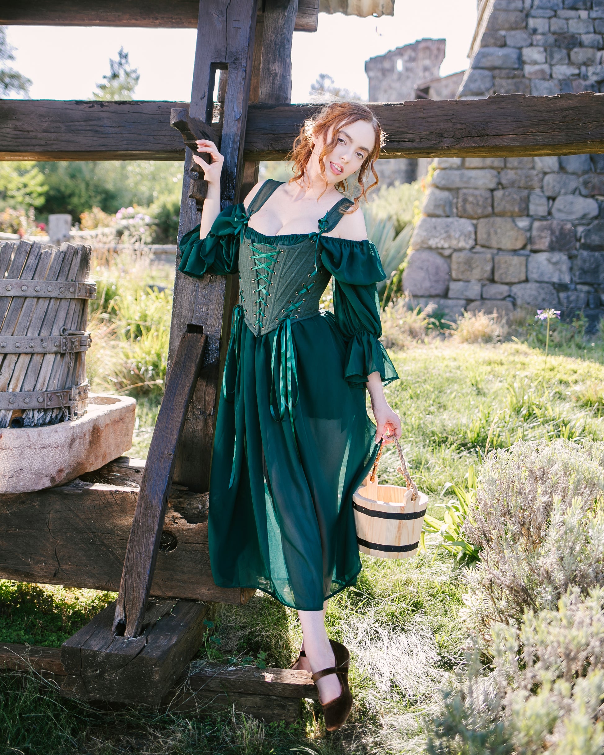 Woman wearing Forest Green Princess Stays corset dress outdoors near rustic wooden structure holding a small basket