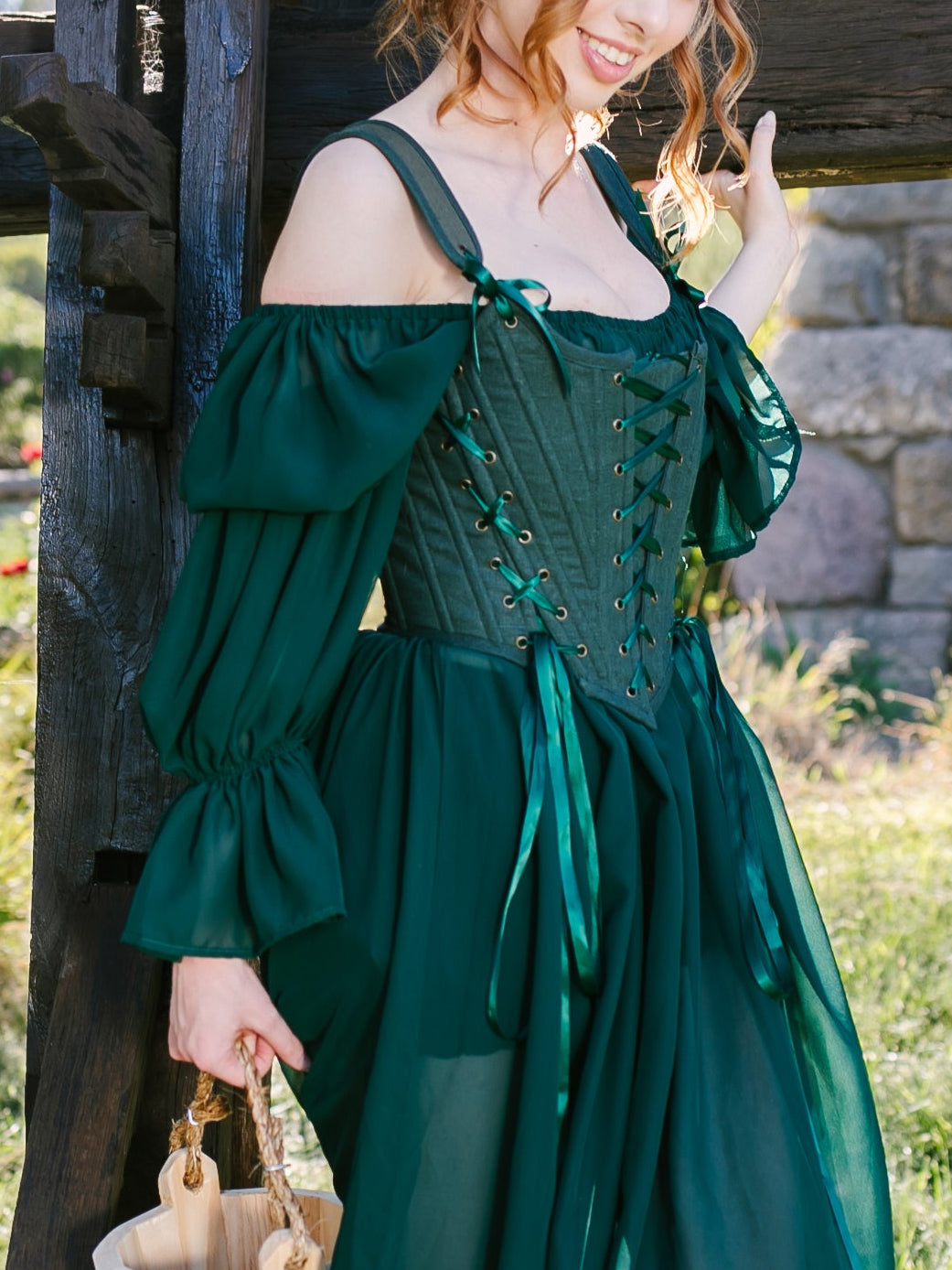 Woman wearing a forest green Princess Stays corset dress outdoors standing by a wooden beam in soft sunlight