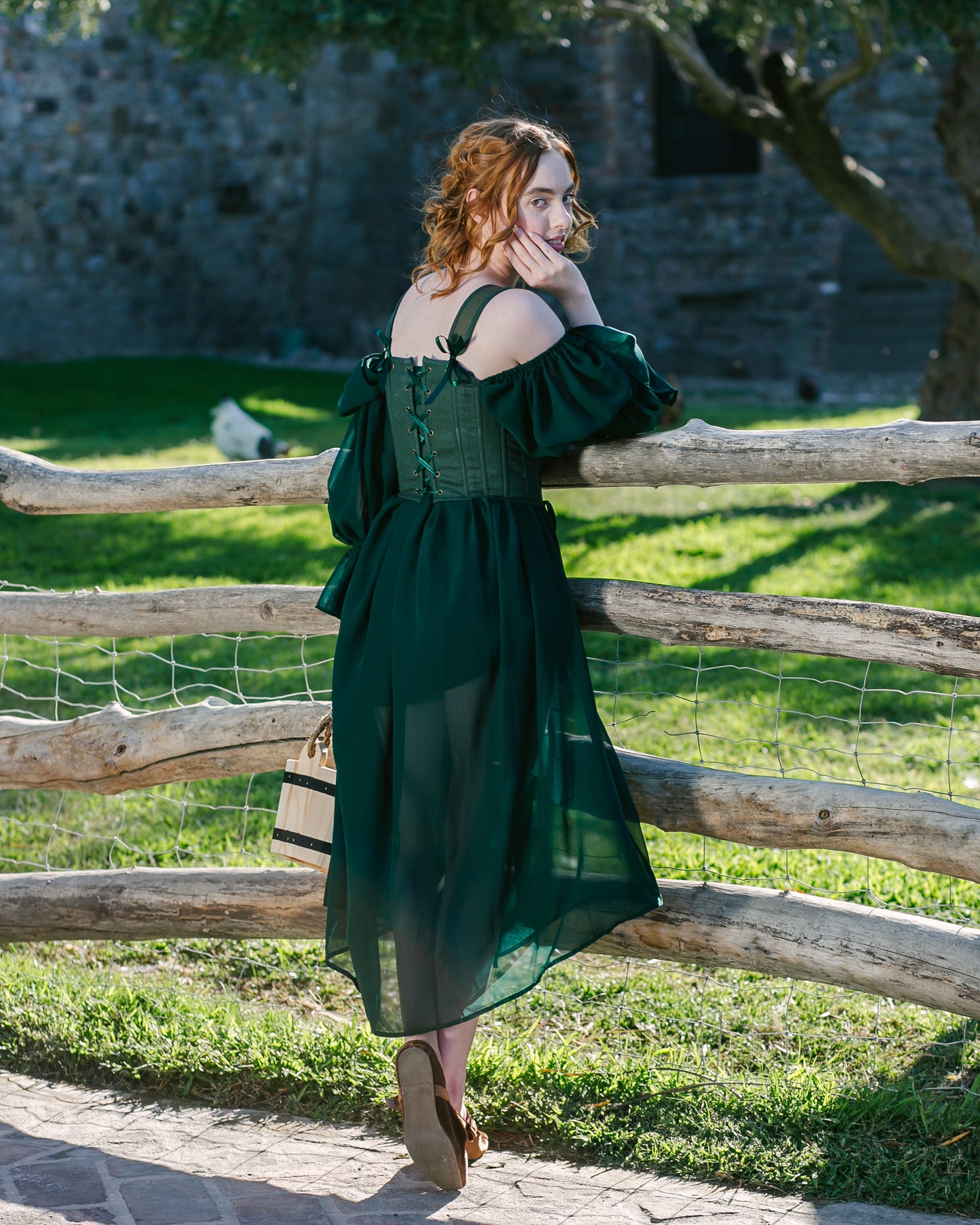 Woman wearing a Forest Green Princess Stays corset with flowing dress standing outdoors by wooden fence in daylight